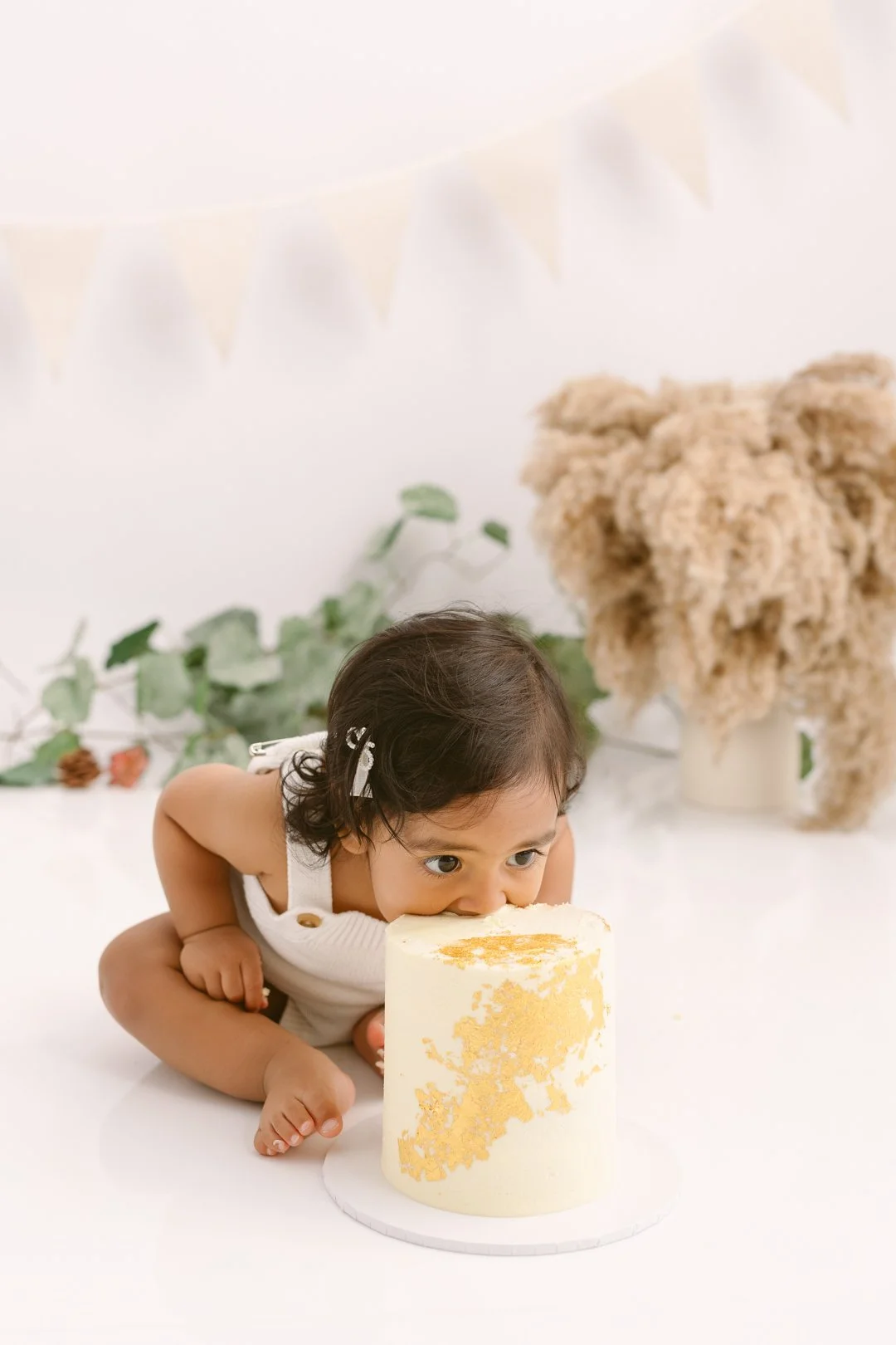 A  one year old baby is biting a decorated yellow and white cake on a white surface with greenery and fluffy decorations in the background.