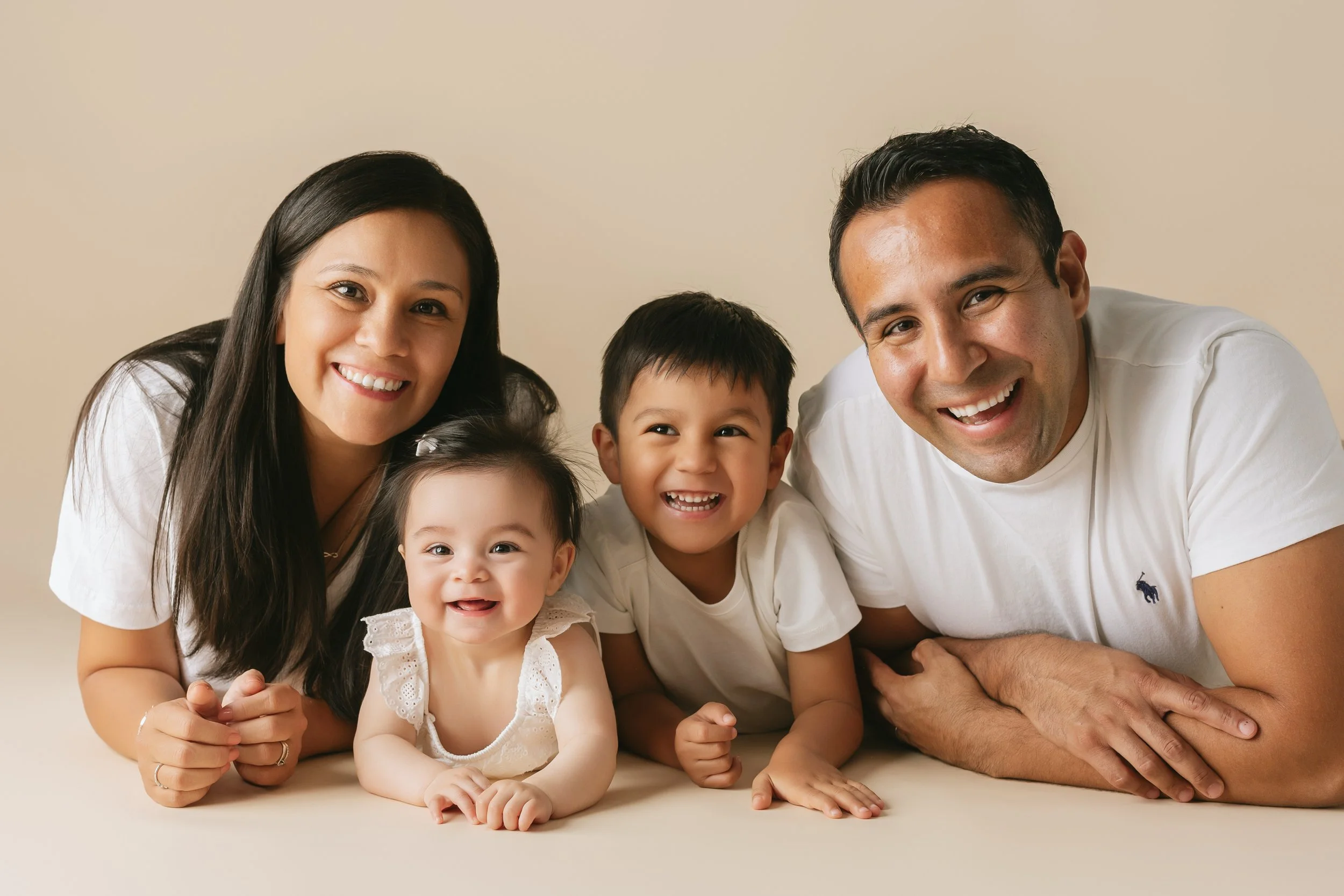 Family of four laying on the floor, smiling at camera, including mum , dad , 4 years old brother, and a toddler girl, all wearing white tops.