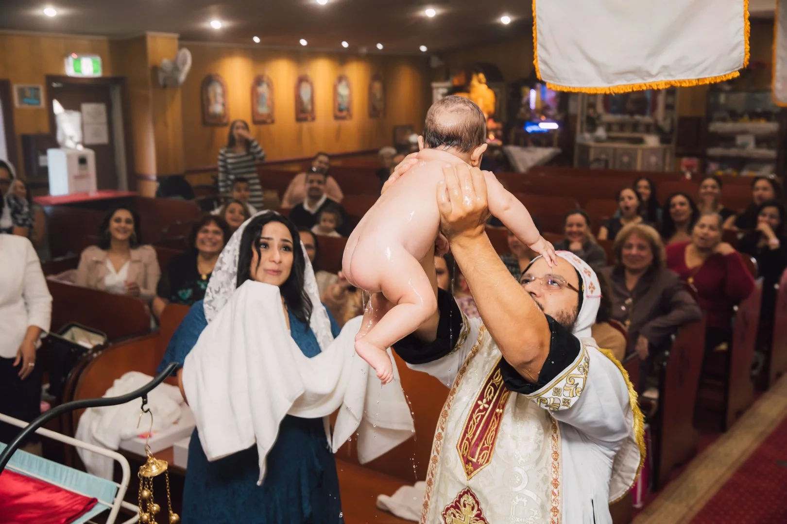 A baptism ceremony taking place in a church with a priest pouring water over a crying baby's head held by a woman, while a woman in a blue dress with a white veil looks on in surprise, and a congregation of smiling people watches in the background.