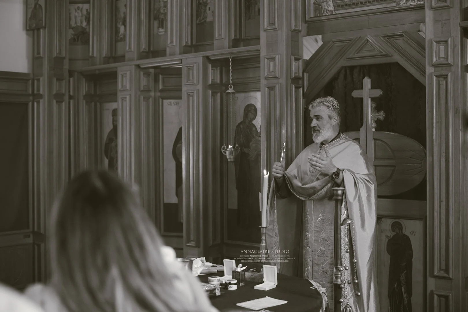 A priest dressed in ornate religious robes conducts a church service inside a wooden church. He holds a candle and gestures with his right hand, while a woman with long hair sits in the foreground. Icons and religious artwork adorn the wooden walls.