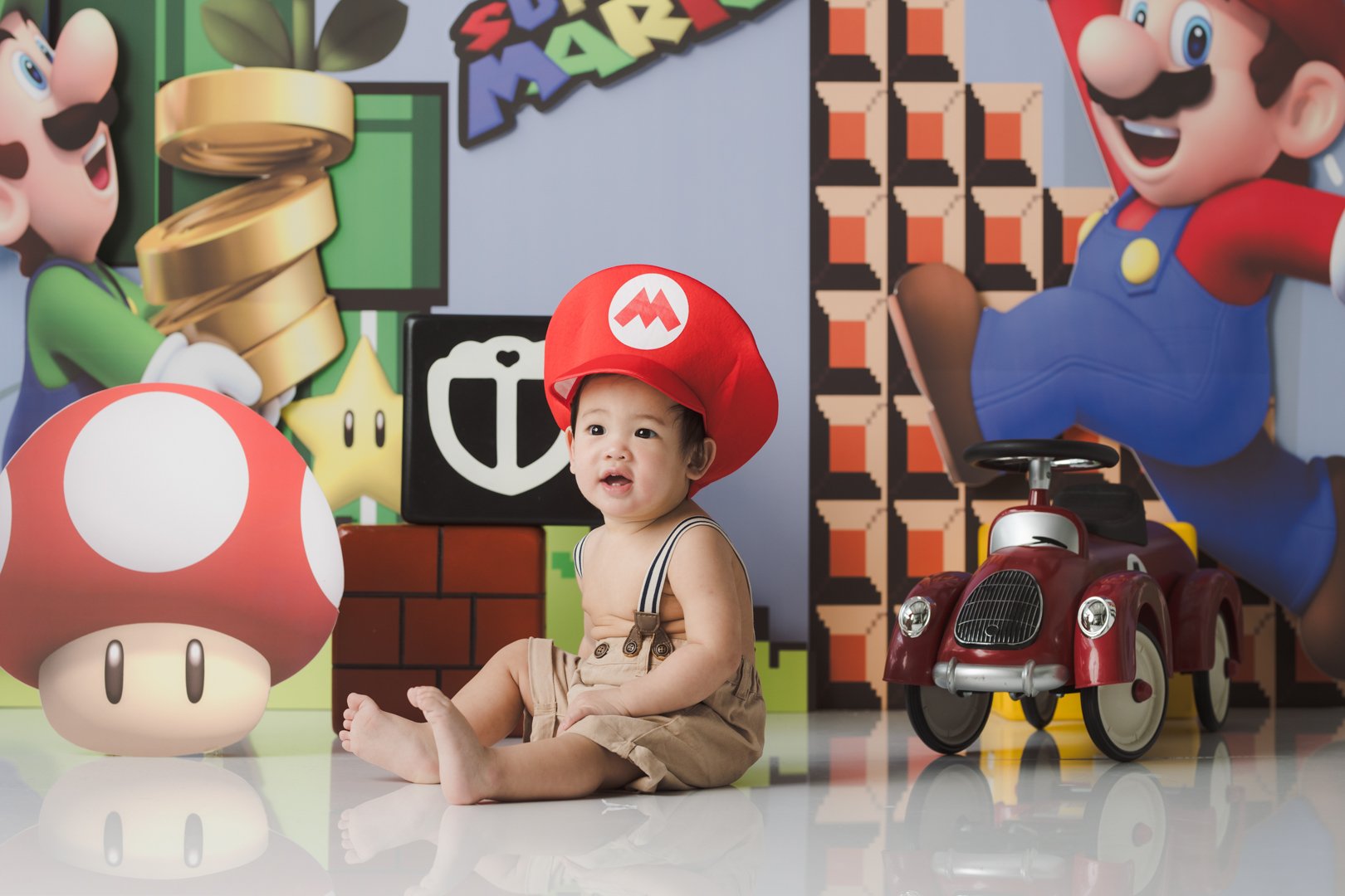  dressed in tan shorts and suspenders, wearing a red Super Mario hat, is sitting on a reflective white floor in front of a Super Mario themed backdrop. The backdrop features characters including Mario and Luigi, along with elements like a Mega Mushro