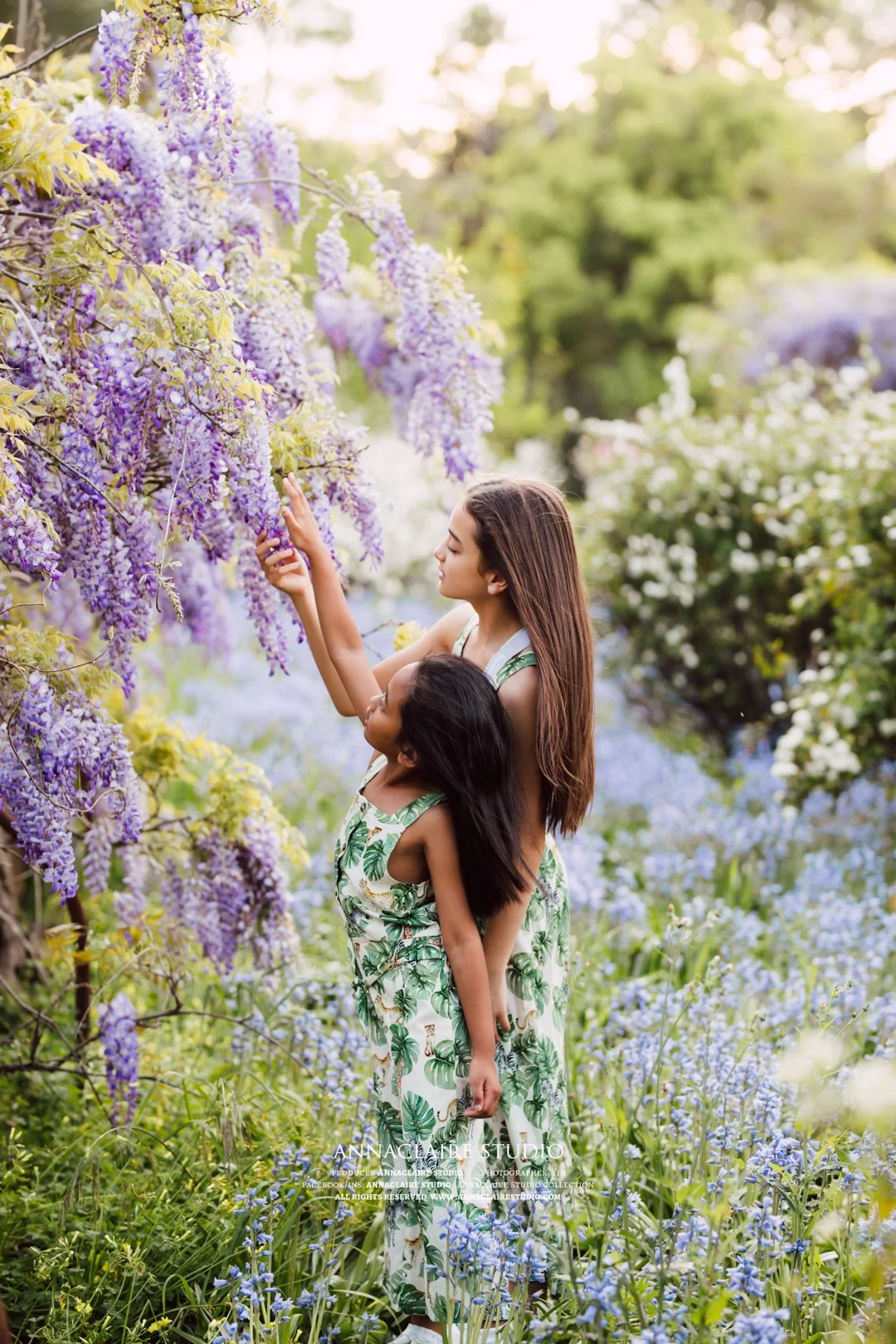 A woman and a young girl are standing in a lush garden with purple and white flowers. The woman is holding a branch of flowering purple wisteria, and the girl is standing close, looking up at the flowers, both dressed in matching green leafy patterne