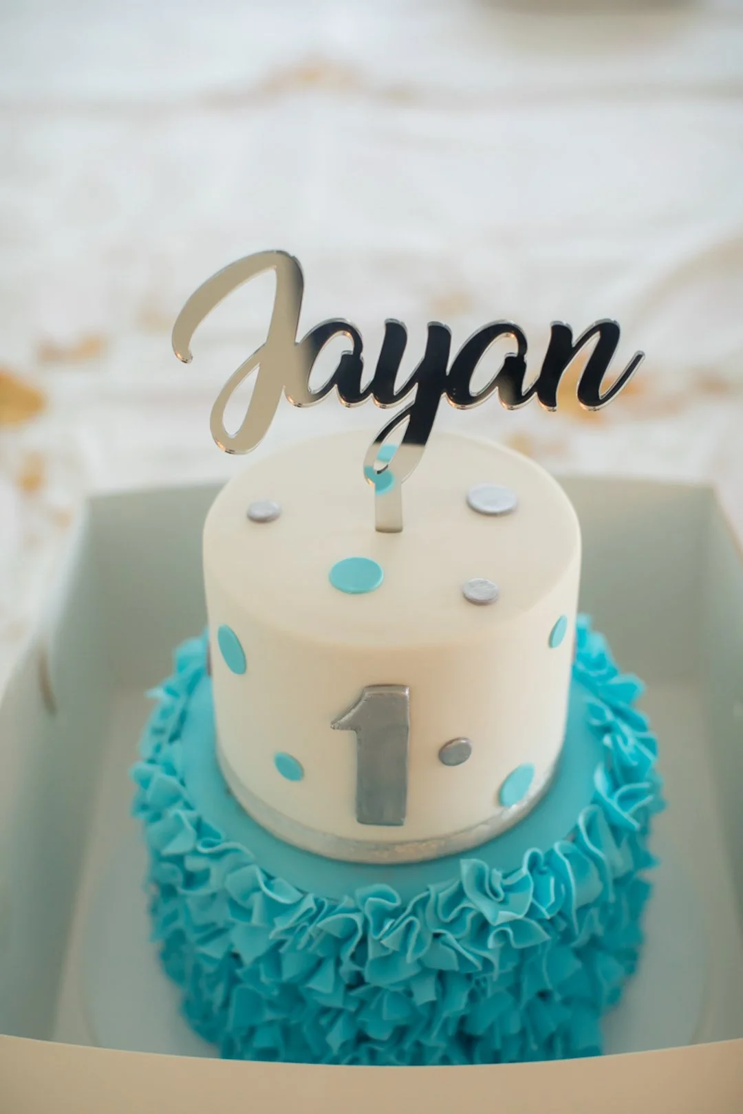 A birthday cake with white and blue decorations, topped with a silver 'Jayden' cake topper and a large silver number one, placed inside a white box.