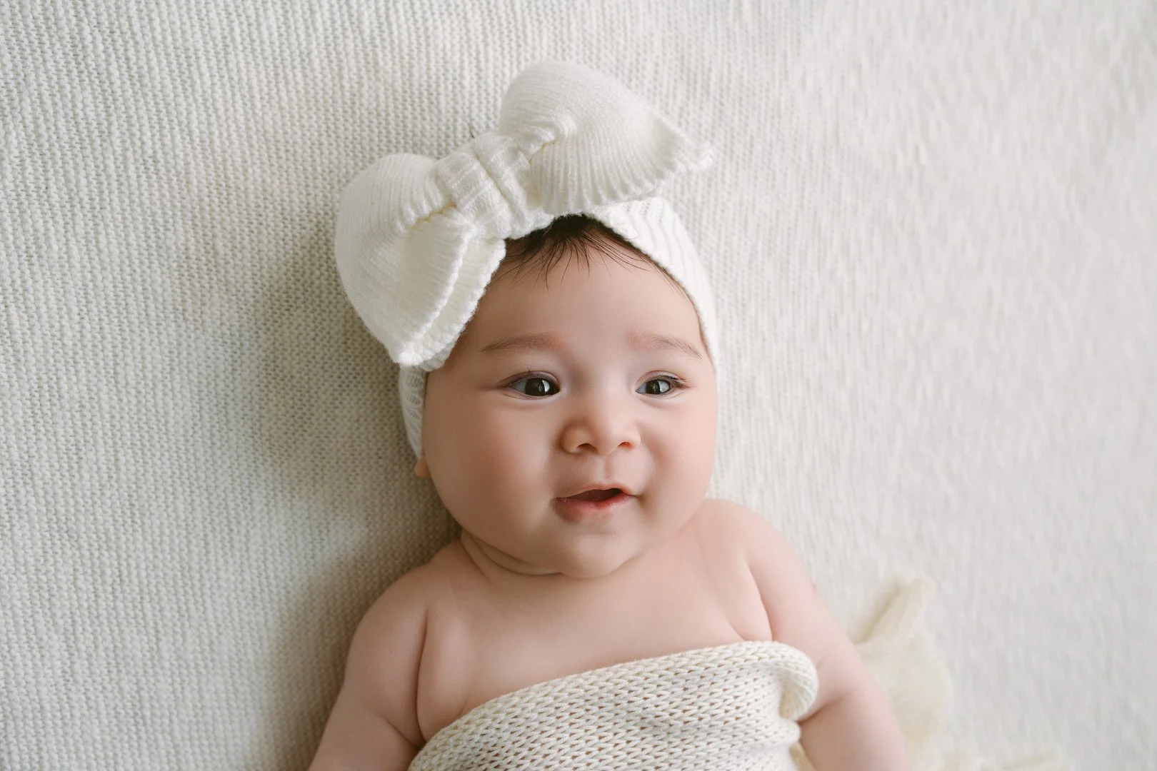 Close-up of a smiling baby with a white headband and bow, lying on a cream-colored textured blanket.