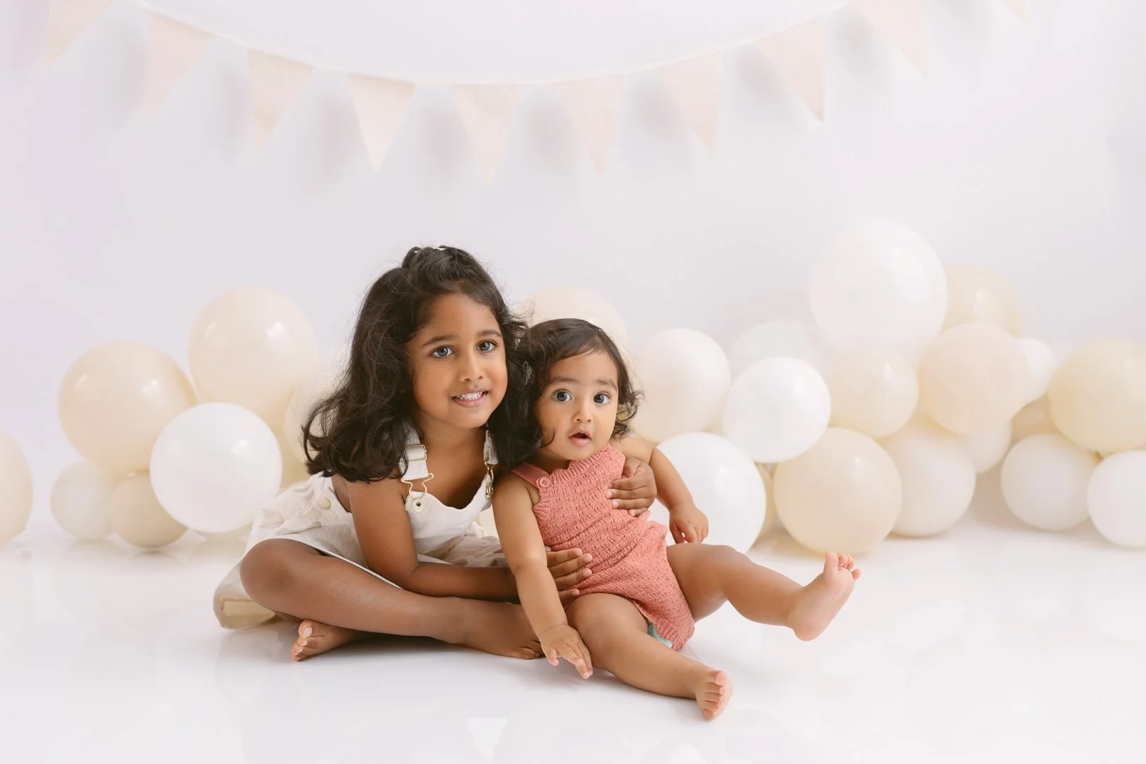 Two sisters sitting on the floor surrounded by white balloons, with a plain white background and string of beige pennant banners hanging above them.