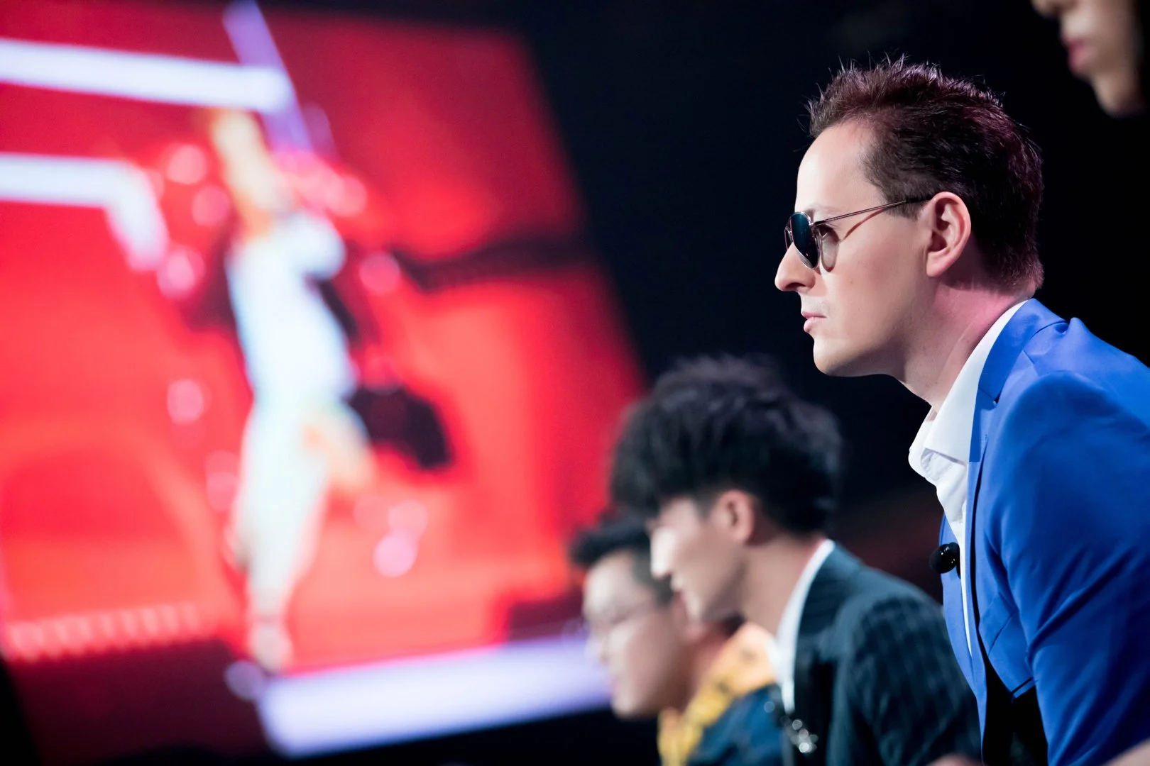 A group of young men sitting at a table, dressed in formal attire, with a man in the foreground wearing sunglasses and a blue suit, watching an electronic chess game on a screen.