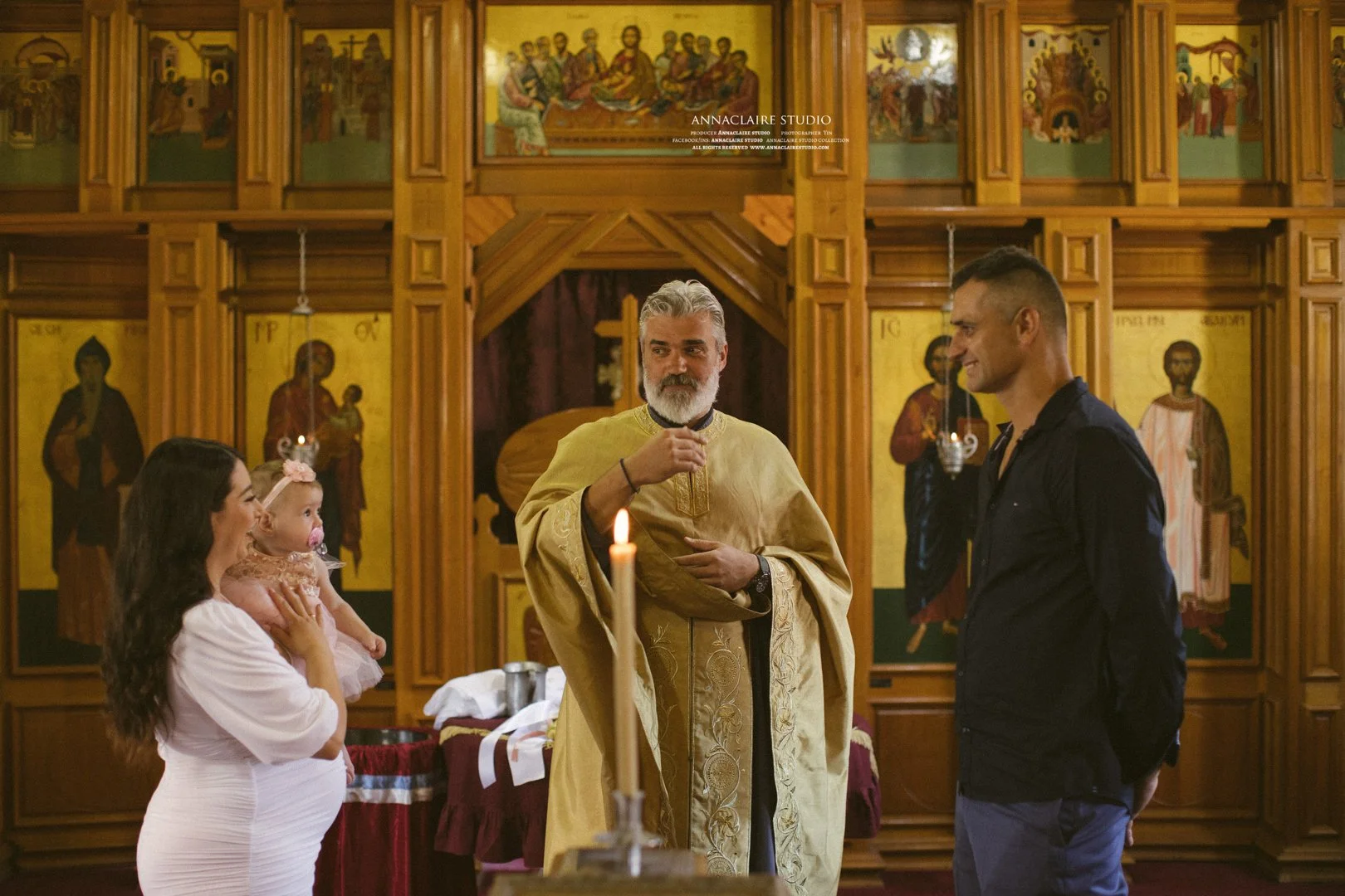 A religious ceremony inside an Orthodox church with icons on the wall. A priest in gold robes is speaking, a woman holds a baby, and a man stands nearby.