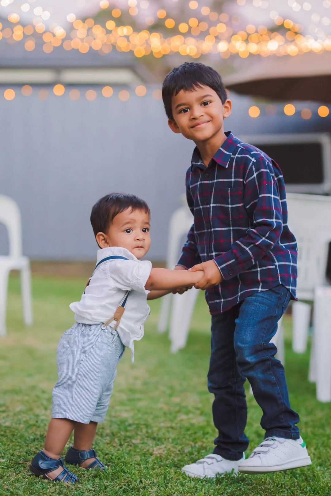 Two young boys are holding hands and dancing together outdoors on a grassy yard. The older boy is smiling and looking at the camera, while the younger boy is looking slightly to the side. Chairs and string lights are visible in the background.
