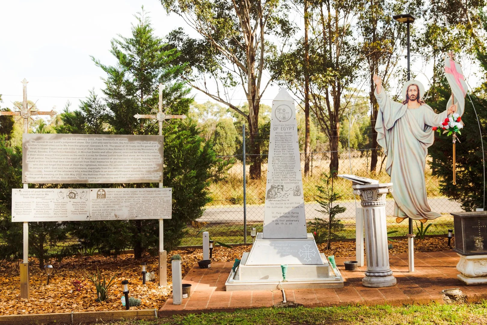 Memorial site with a statue of Jesus Christ with open arms, a cross with flowers, informational plaques, and trees in the background.