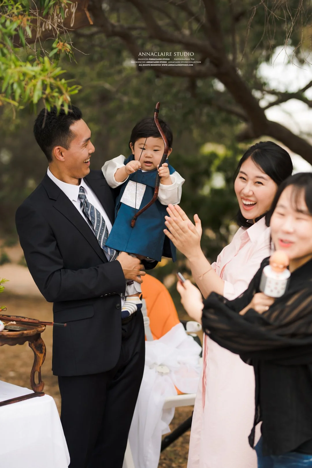 A man holding a young child dressed in traditional Korean attire, smiling as another woman claps and laughs nearby, in an outdoor setting with trees.