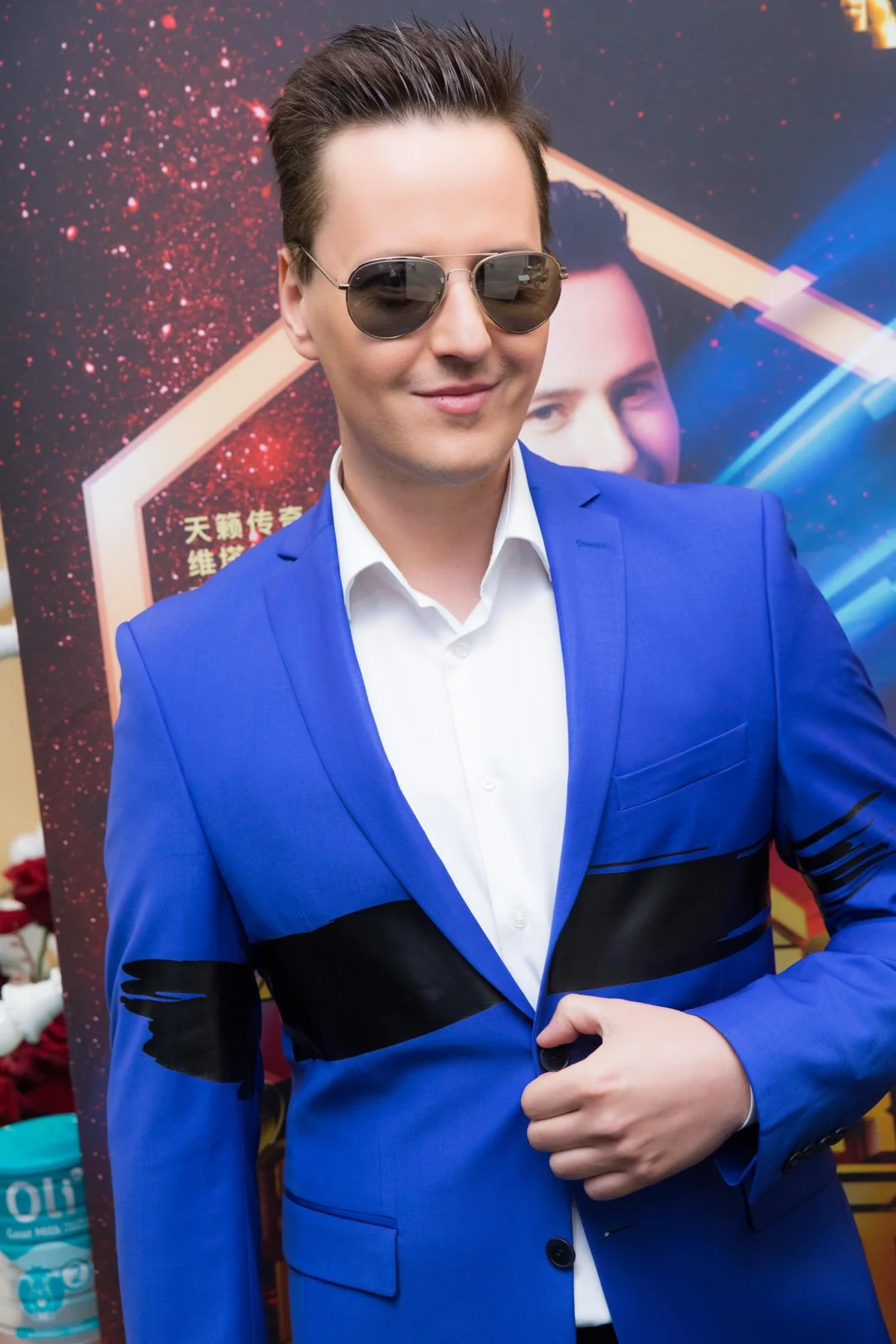 A man is wearing a white shirt, a blue blazer with black accents, and sunglasses. He has short, styled hair and is smiling. Behind him, there is a colorful background with a large poster featuring a cartoon or animated character and some text.