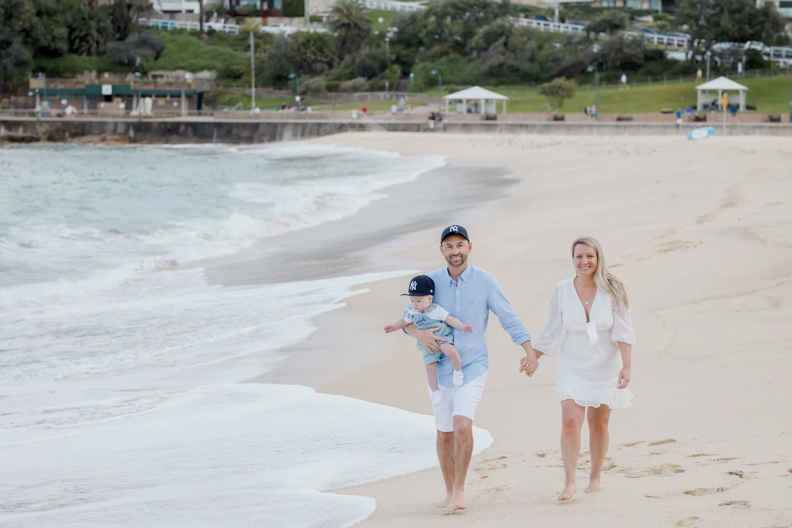A family of three walking hand in hand along the beach near the shoreline, with waves and a green park in the background.