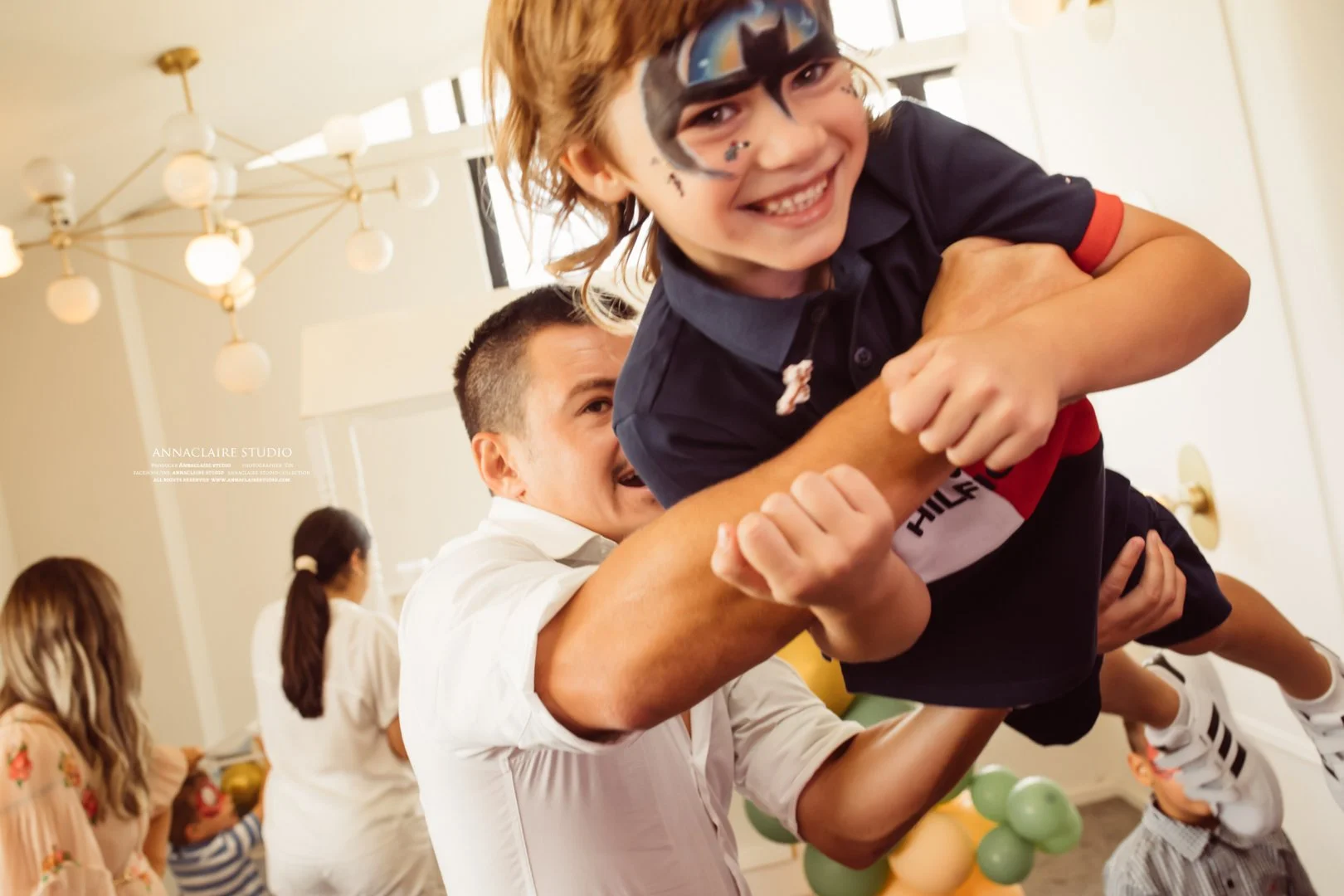 A young boy with face paint smiling as he is lifted and playfully wrestling with an adult man, in a room decorated for a celebration with balloons.