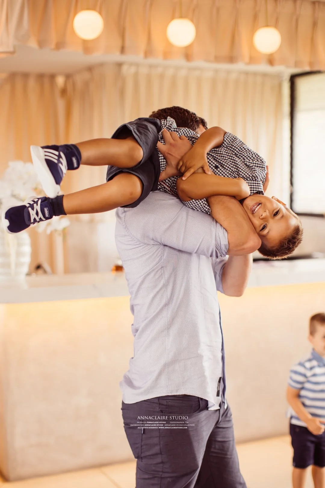 A man lifting a smiling young boy in the air indoors with warm lighting, wooden walls, and another child in the background.