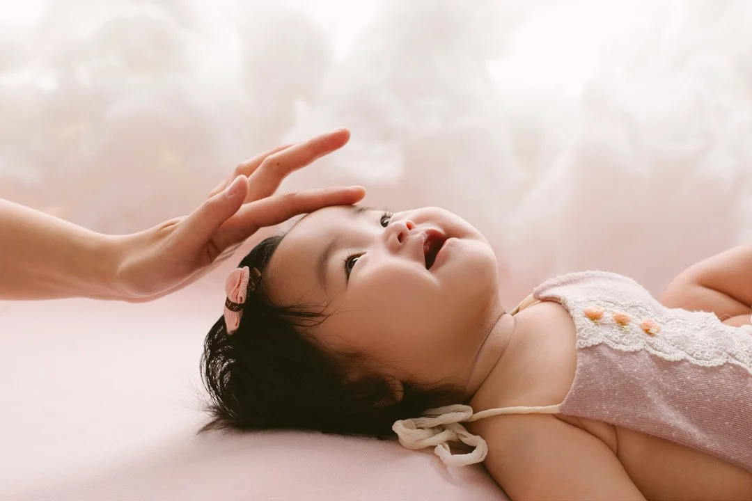 A young child with dark hair lying on a pink surface, looking up and smiling while an adult gently touches her forehead.