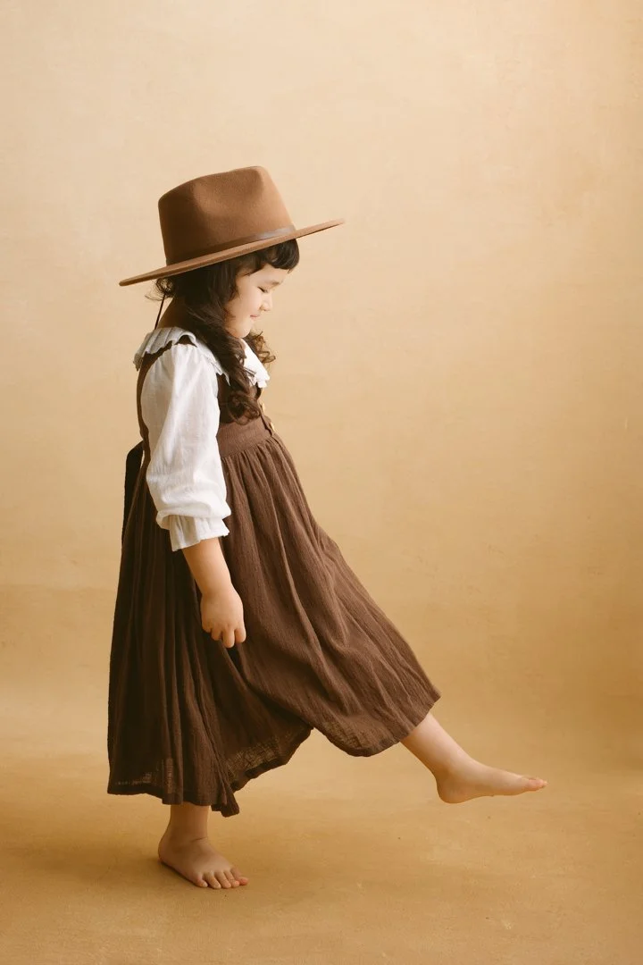 A young girl wearing a brown wide-brimmed hat, white long-sleeve blouse, and a brown jumper dress, standing barefoot against a beige background. She is lifting her right foot slightly off the ground, looking down with a gentle smile.
