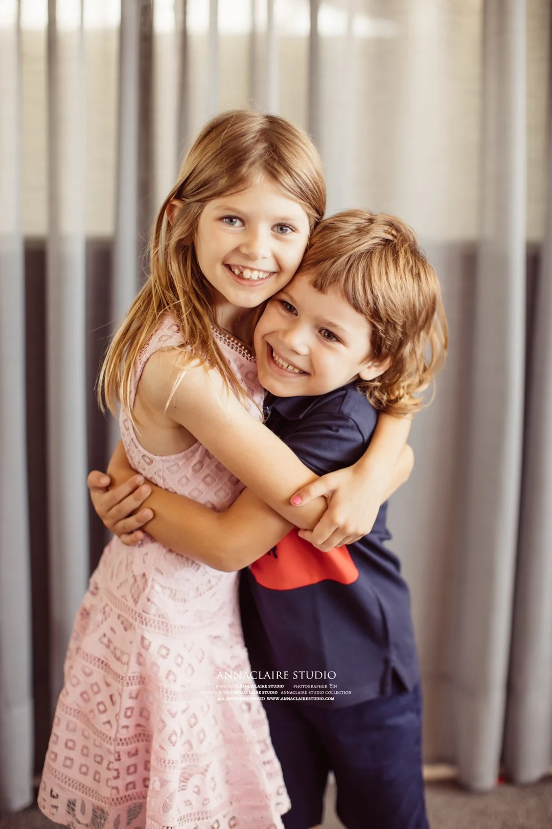 Two children, a girl and a boy, hugging each other and smiling in front of a curtain.