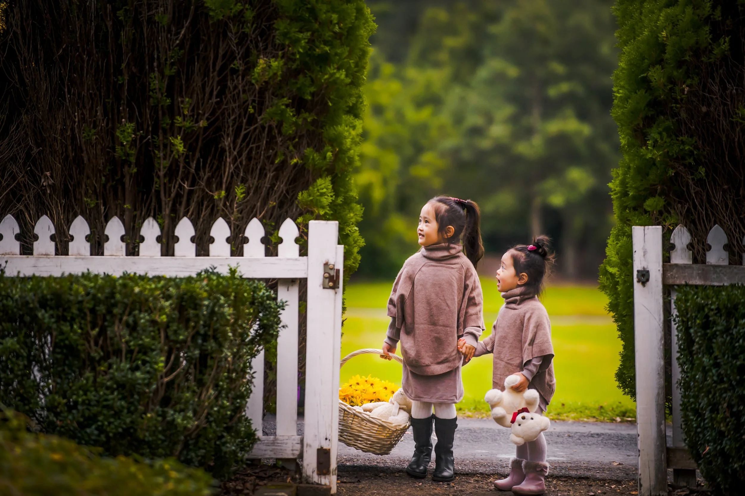 Young sisters holding hands in a garden gate, with one carrying a basket of flowers and the other holding a stuffed animal.