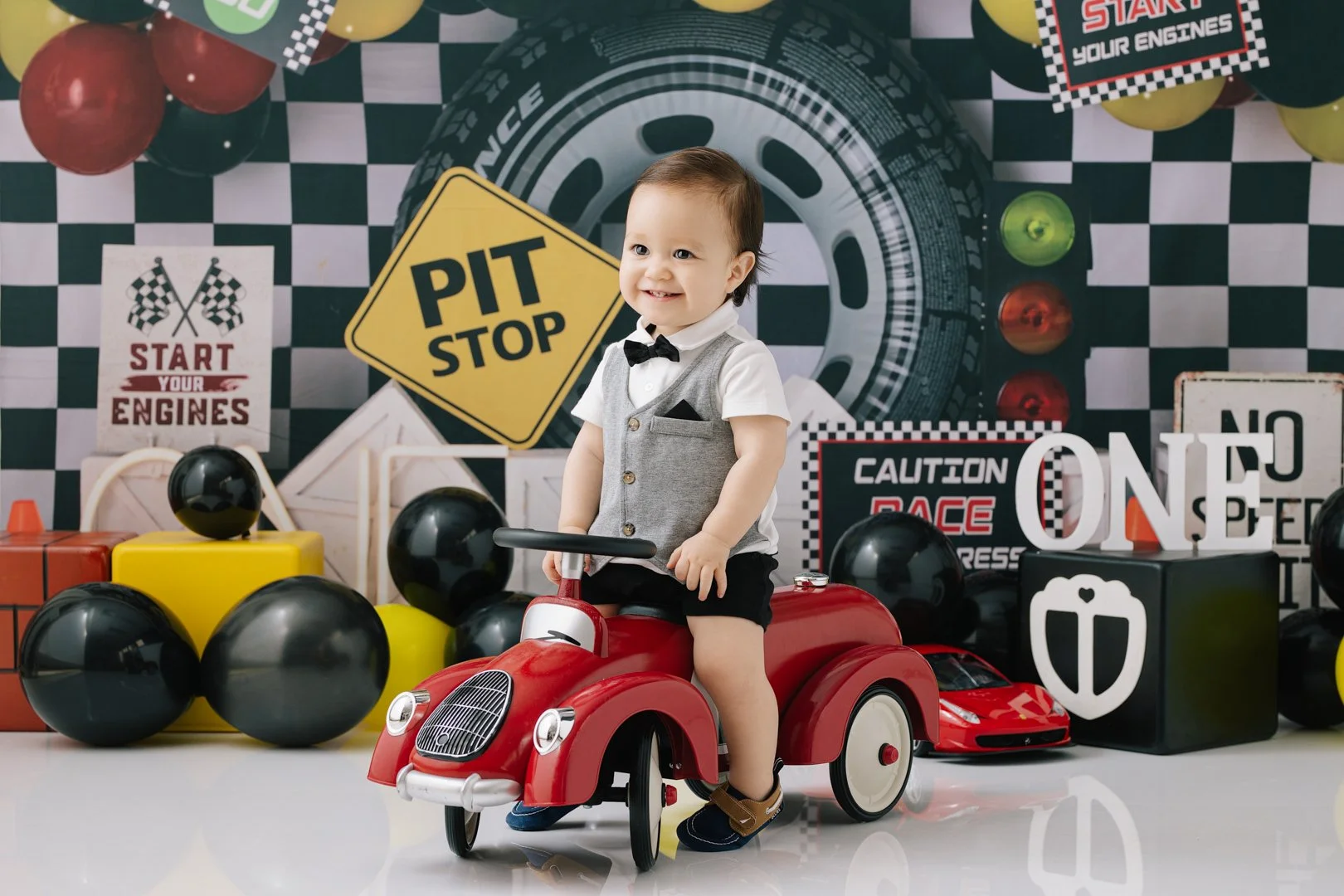 A  one year old baby boy dressed in a vest, bow tie, and shorts, sitting on a red toy car, smiling in front of a racing-themed backdrop with racing signs and decorations.