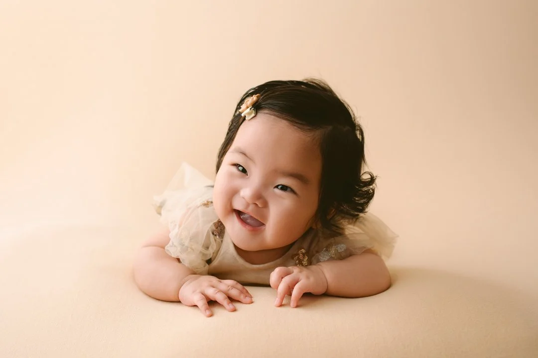 A smiling baby girl lying on a neutral-colored surface, wearing a light-colored dress with tulle details and a small bow in her hair.