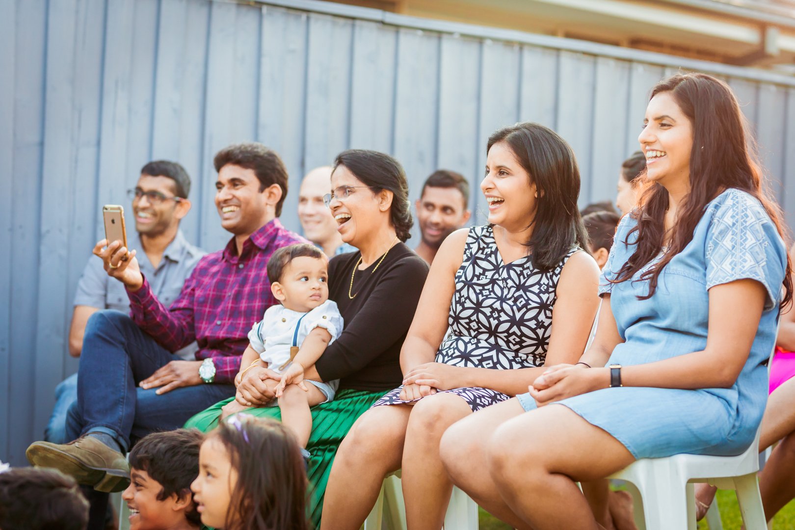 Group of people seated outdoors, smiling and laughing, with a blue fence in the background.