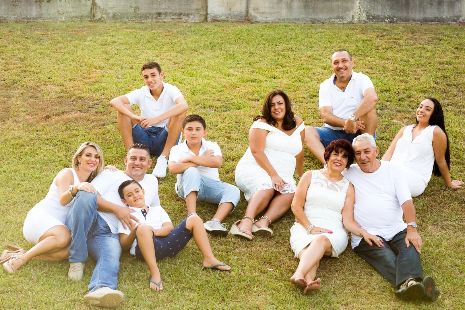 Family group sitting and lying on grass in a backyard, smiling, with a stone wall in the background.