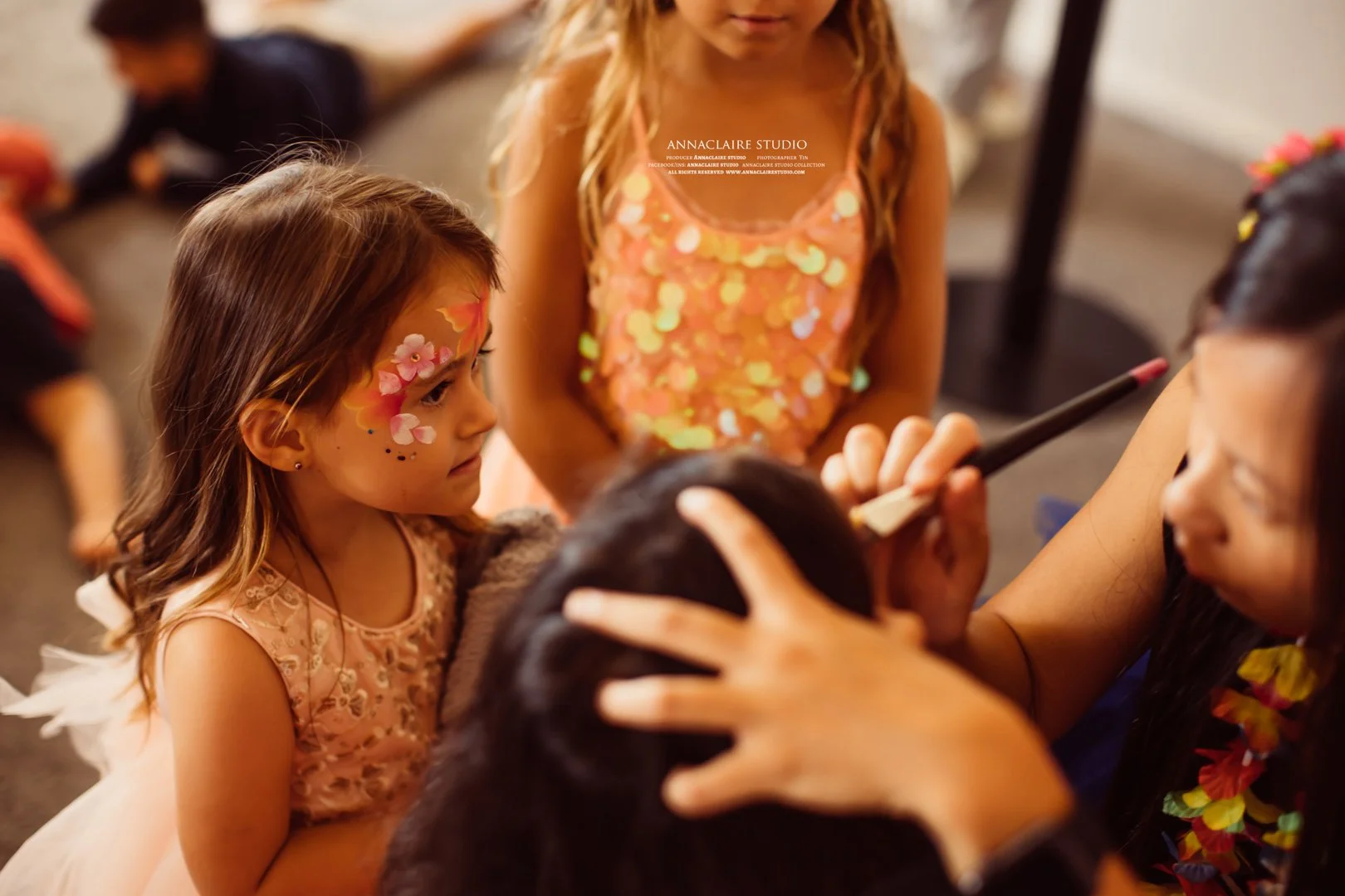 A face painter is applying pink and white floral face paint to a young girl with light brown hair. The girl is sitting still, wearing a light pink dress with lace details. Other children are in the background, some sitting and some lying on the floor