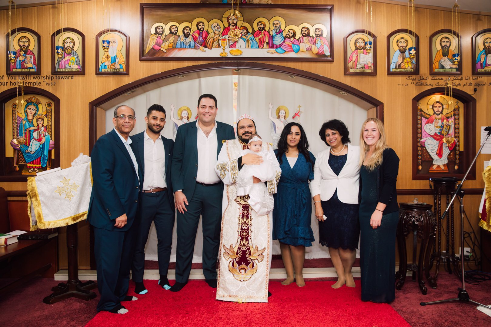 A group of nine people standing inside a church, with a priest holding a baby in the center. The background features religious icons and artwork. The group appears to be celebrating a religious ceremony or baptism.