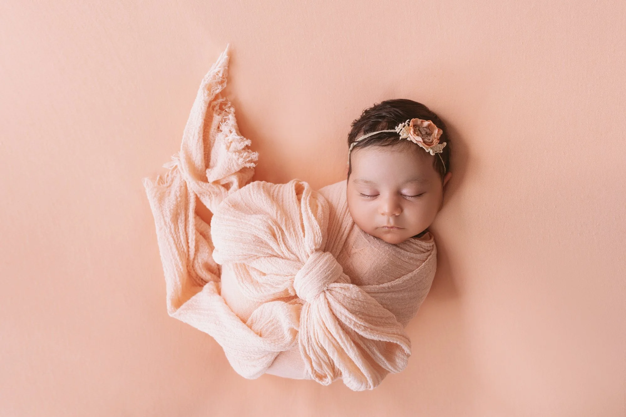A newborn baby girl is sleeping on a peach-colored background, wrapped in a soft pink blanket, with a matching headband that has a flower detail.