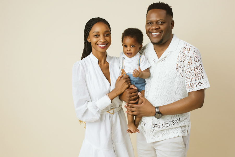 Smiling family of three, a woman, a man, and a baby, posing together against a beige background.