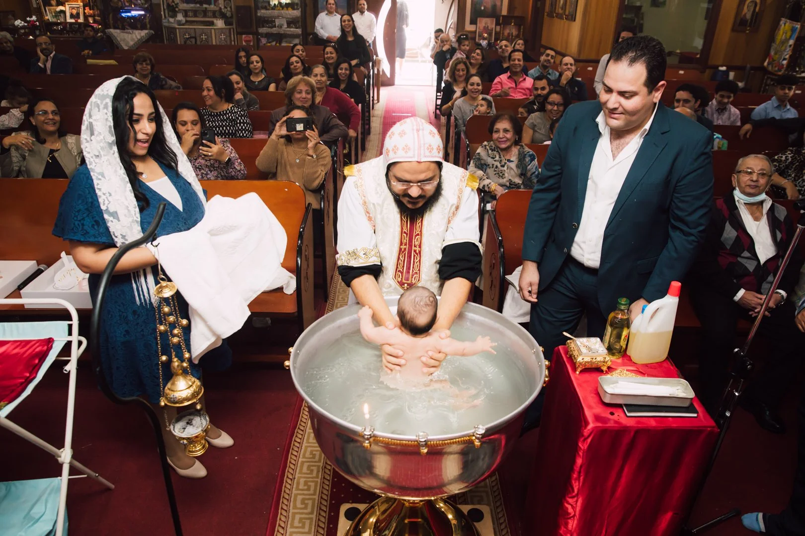 A baby being baptized in a church, with a priest holding the baby in a baptismal font, surrounded by family and friends. The priest is in traditional attire, and people are smiling and taking photos.