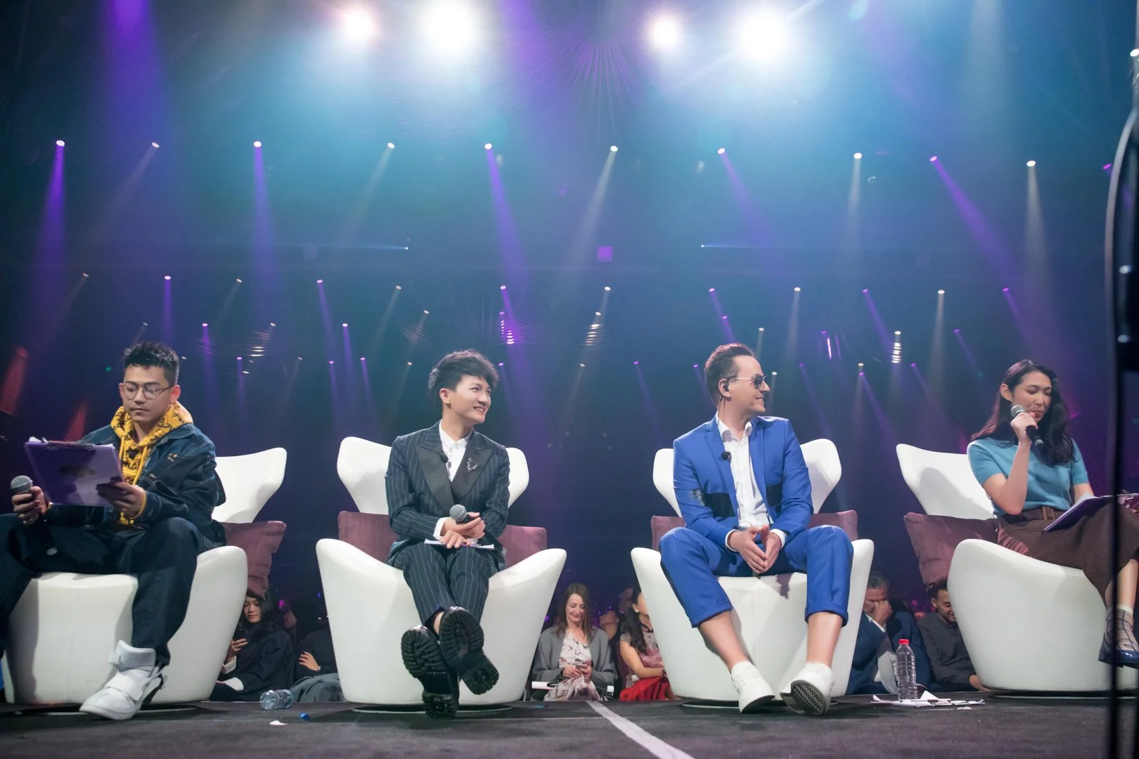 A panel of four people seated on stage under colorful lights, with three women and one man. The man is wearing a blue suit and sunglasses, the woman to his right is in a pinstripe suit, and the woman to his left is in a blue shirt. The person on the 