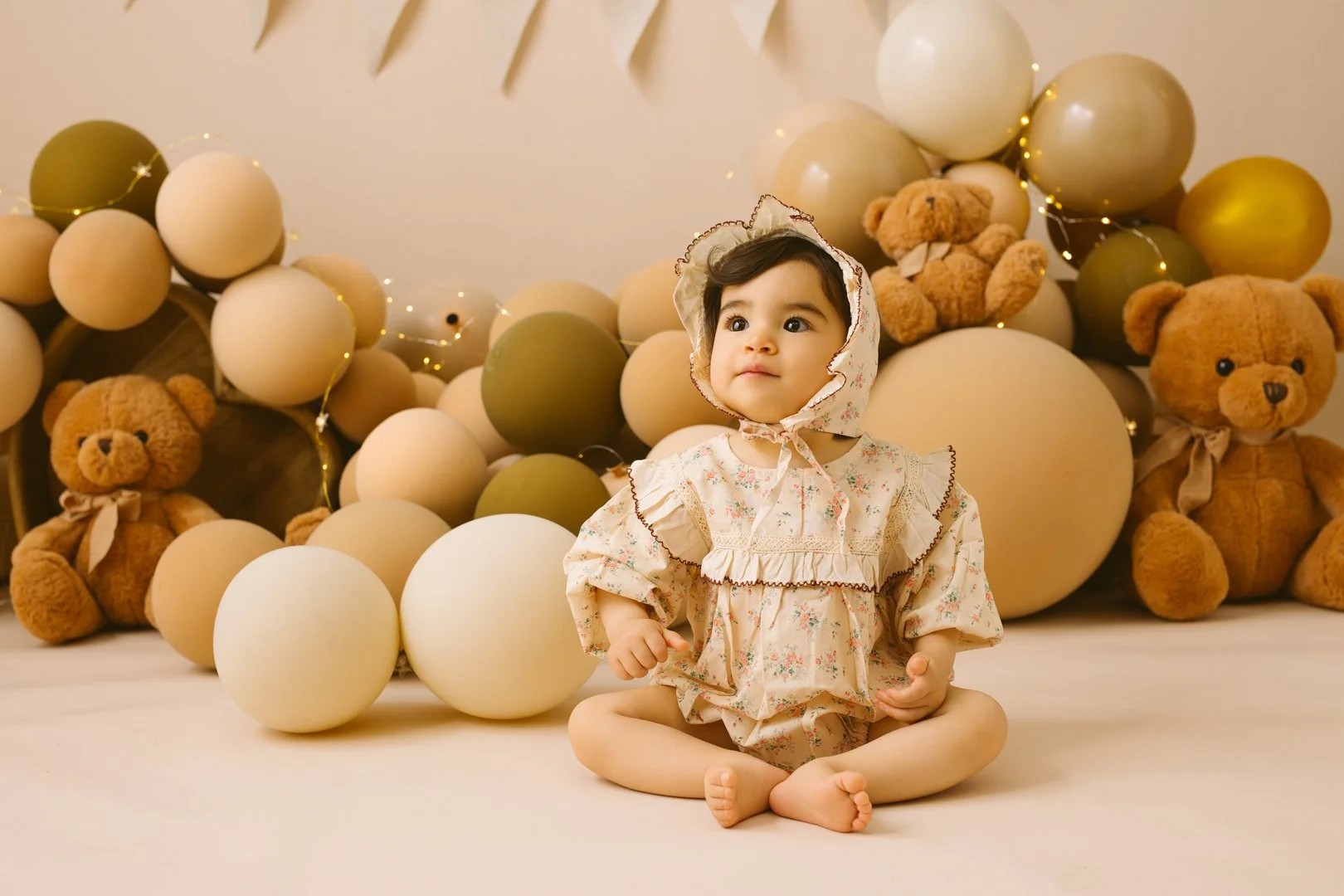 A young girl in vintage floral dress and bonnet sitting barefoot on the floor with a cake smash photo backdrop of large beige, cream, and olive green balloons and teddy bears.