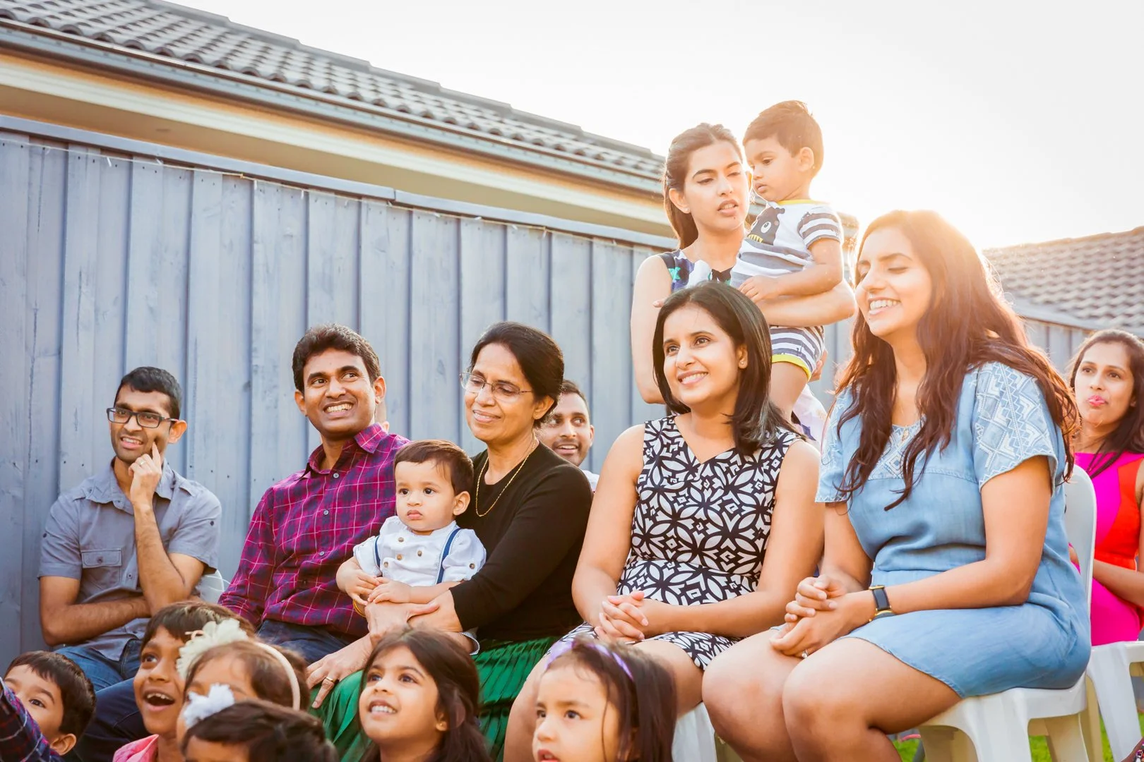 Family and friends gathered outdoors, sitting and standing in front of a blue wooden fence, celebrating and smiling during a sunny day.