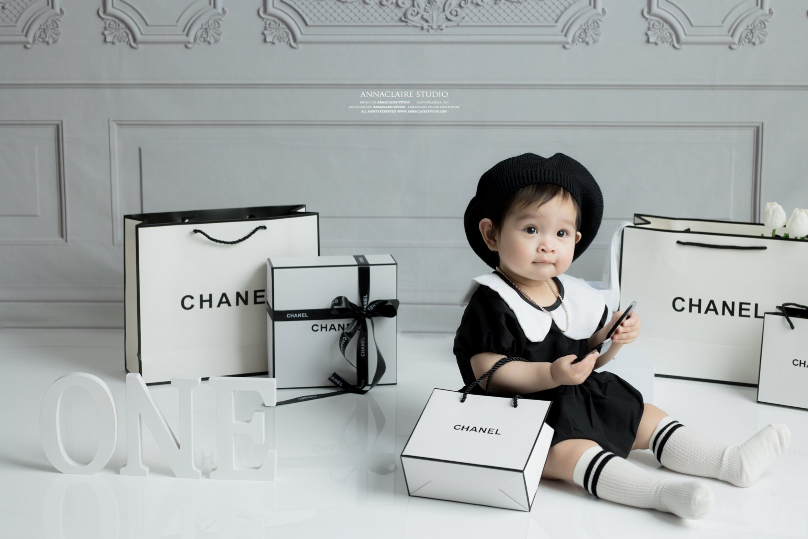 1 year old baby girl  dressed in black and white clothing, wearing a black beret and knee-high socks, sitting on the floor surrounded by Chanel shopping bags and boxes, holding a phone, with decorative molding on the wall behind.