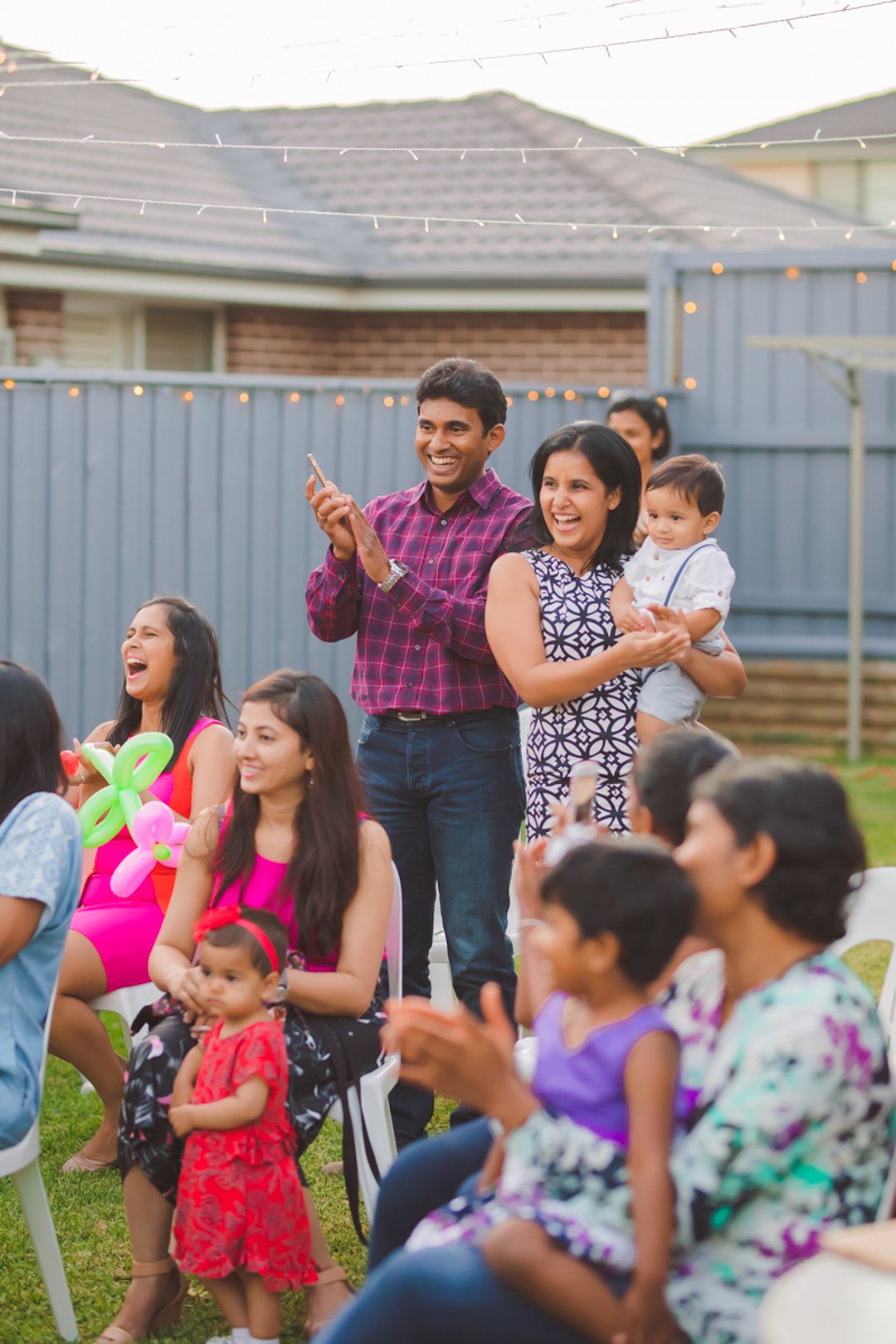 A group of people at an outdoor gathering, smiling and enjoying the event. Some are seated, while others stand. The background shows a fence, a house, and string lights.