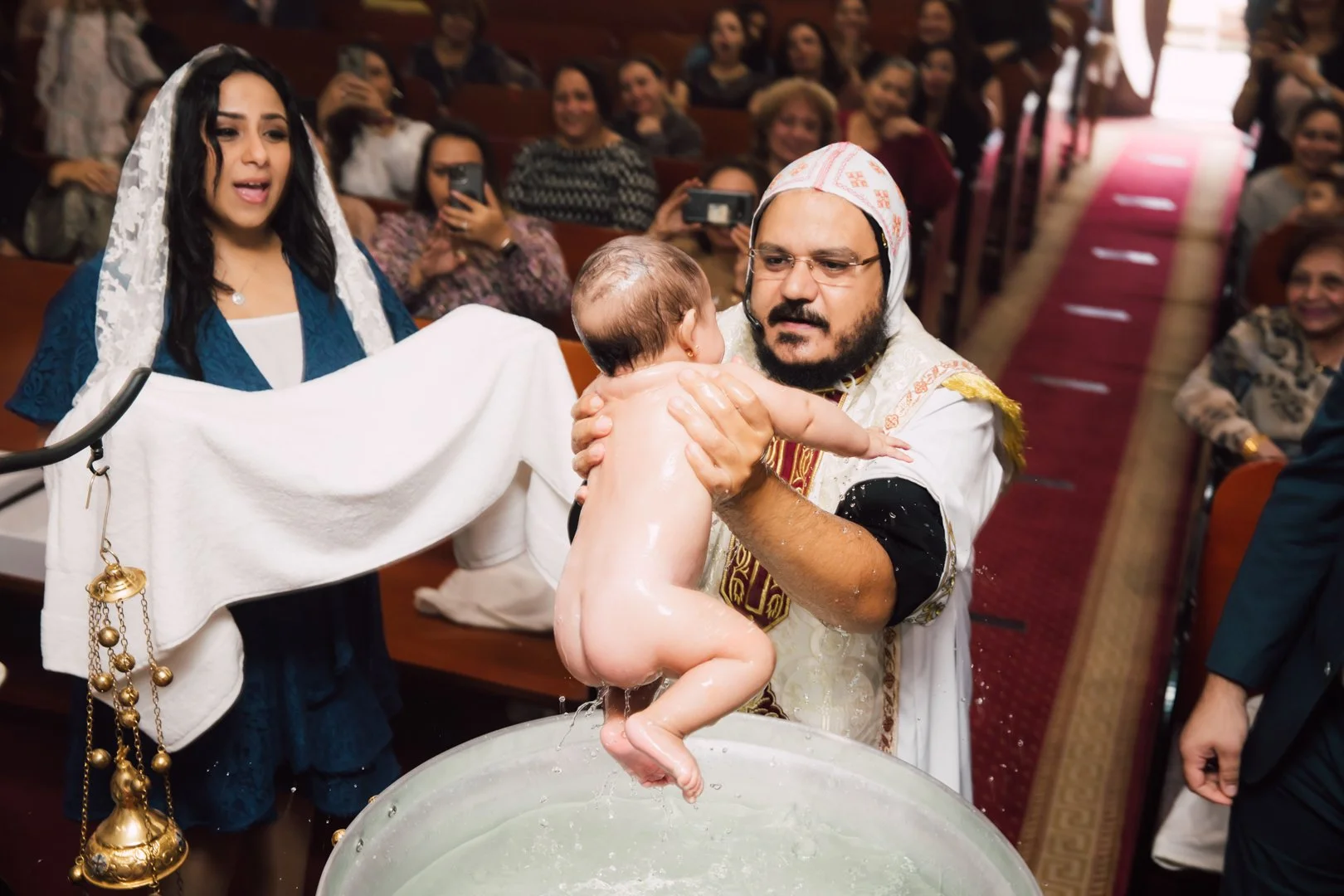 A religious baptism ceremony with a man holding a naked baby over a baptismal font. The woman standing nearby is holding a towel. Many people are watching and some are taking photos, in a church setting.