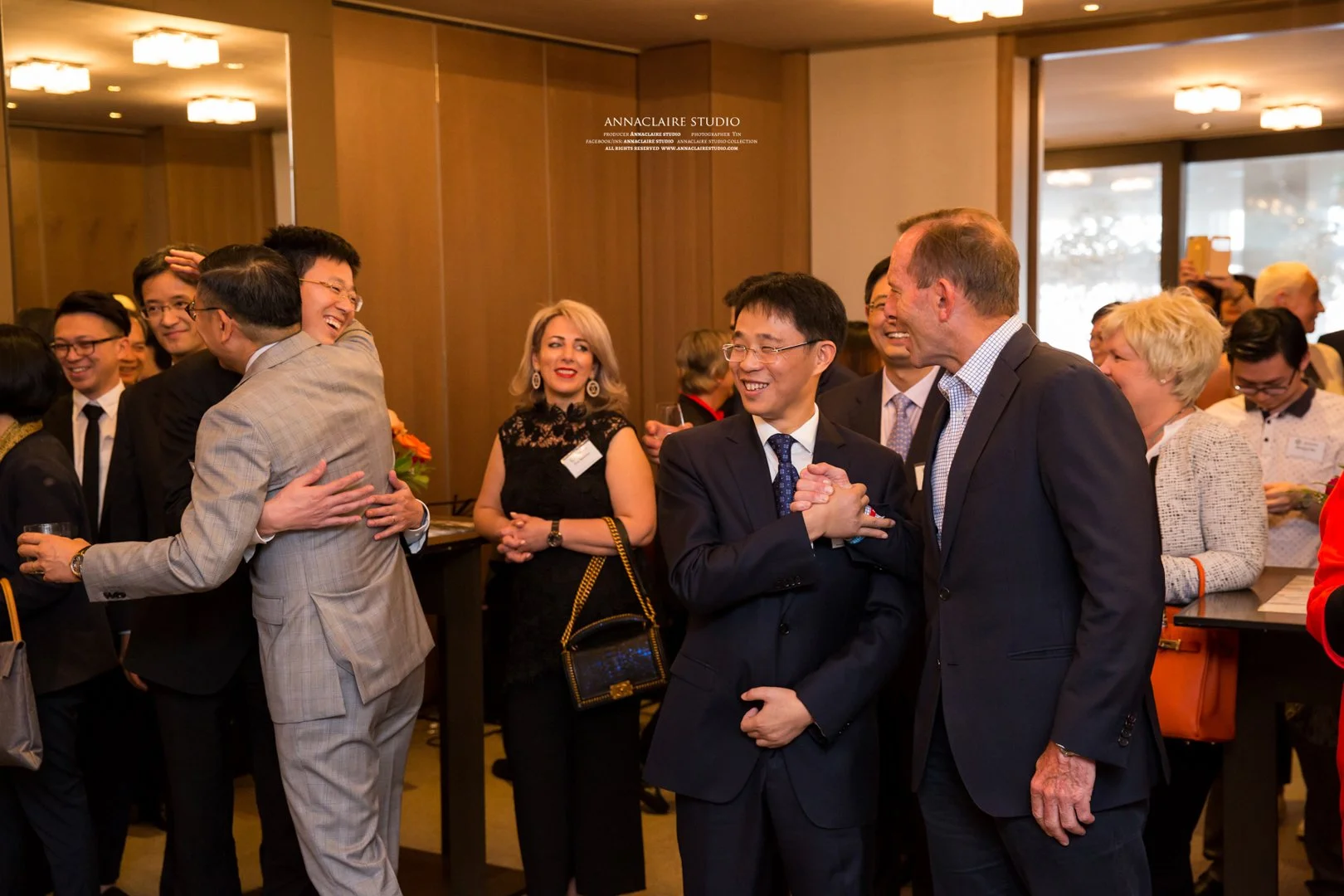 A group of diverse business professionals at a networking event, engaging in conversations, hugging, and smiling in a well-lit, modern conference room.
