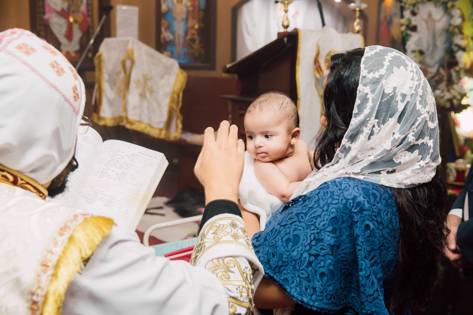 A woman wearing a lace veil holding a baby during a baptism ceremony in a church, with a priest reading from a book.