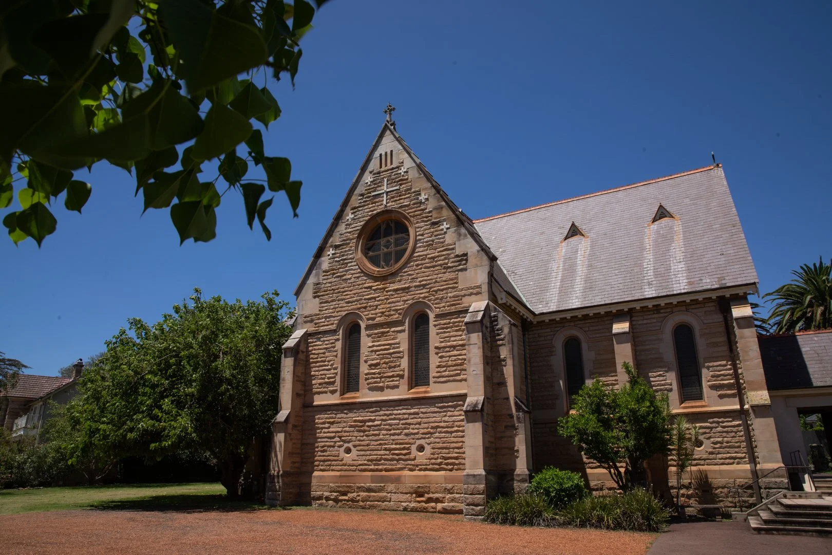 An old stone church building with a sloped roof, arched windows, and a circular window, surrounded by green trees and plants under a clear blue sky.