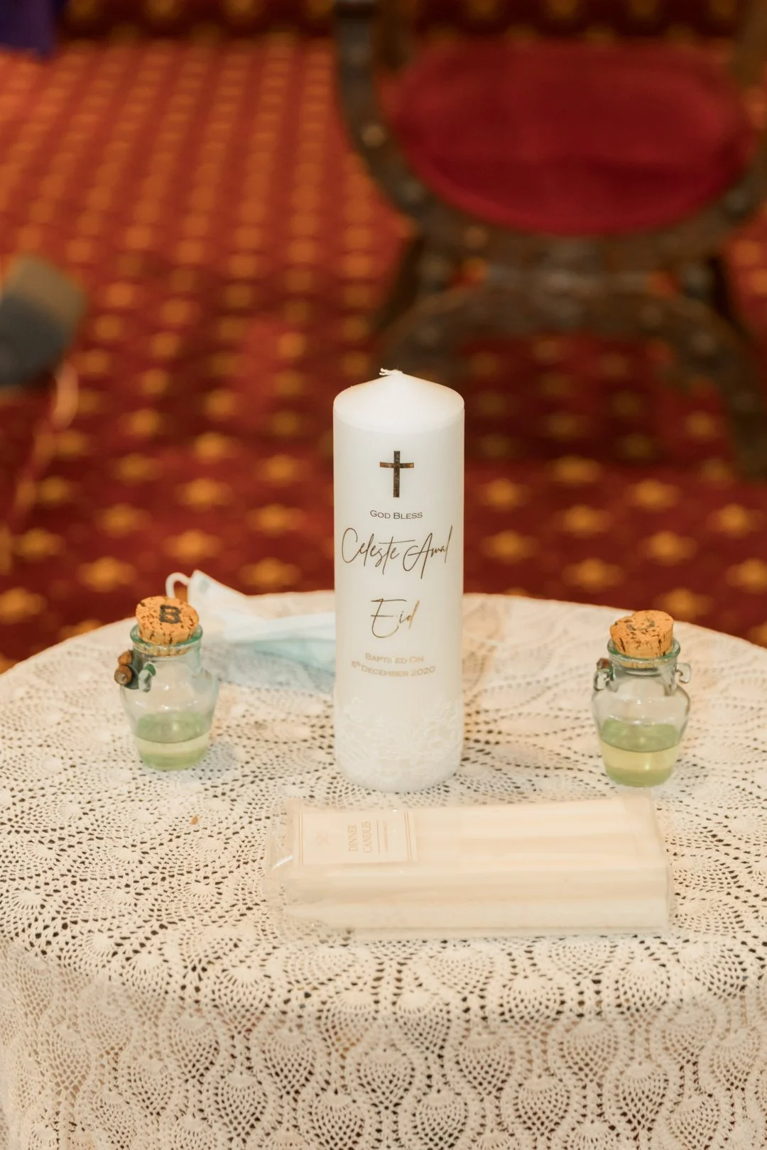 A table with a large white candle labeled 'Celeste Arnel Eid' and two small jars with cork lids containing green liquid, placed on a white lace tablecloth. The candle has a cross symbol and the words 'God Bless' and 'Baptized on December 2020'. A fac