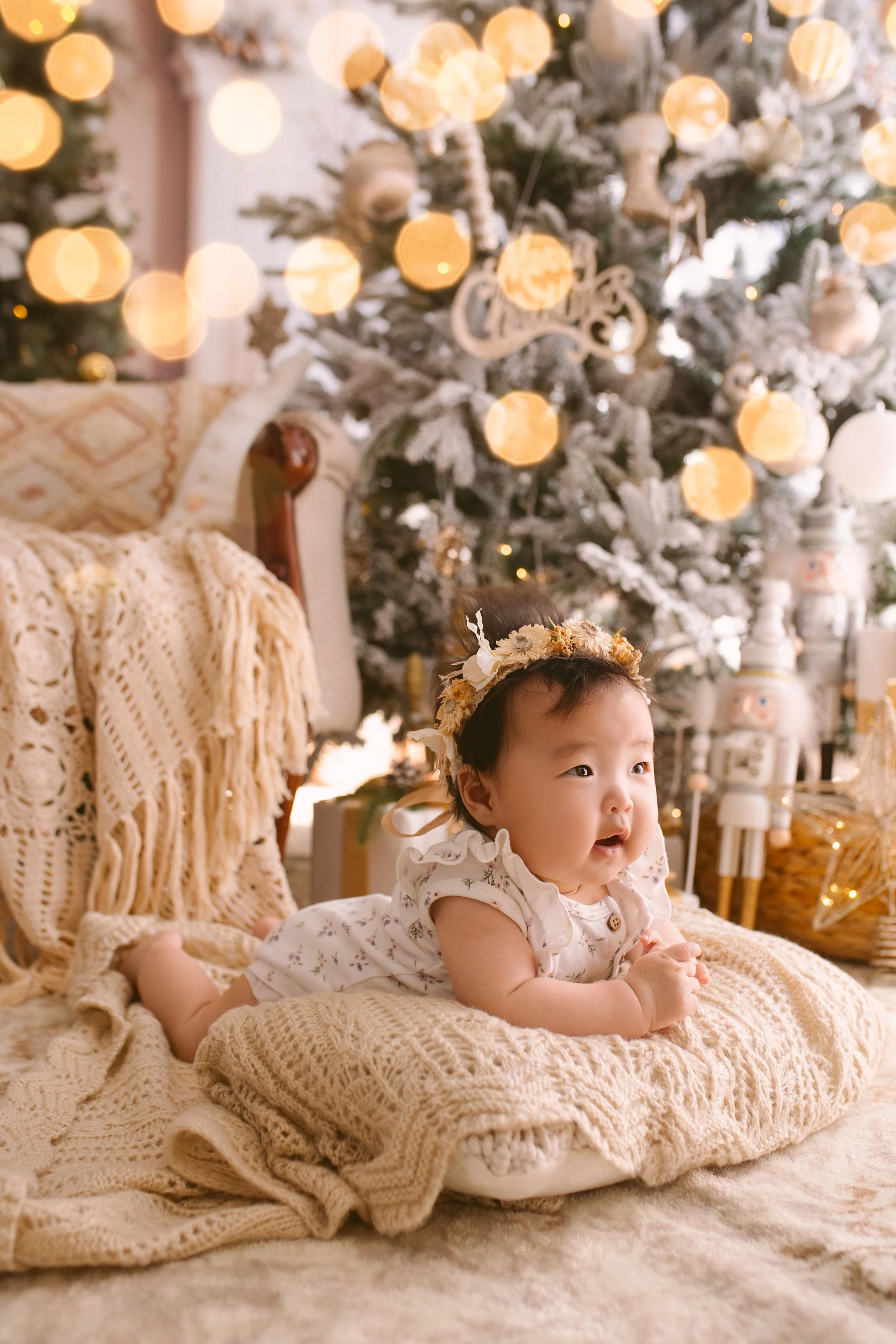 A baby girl with a flower crown lying on a knitted blanket in front of a decorated Christmas tree with lights and ornaments.