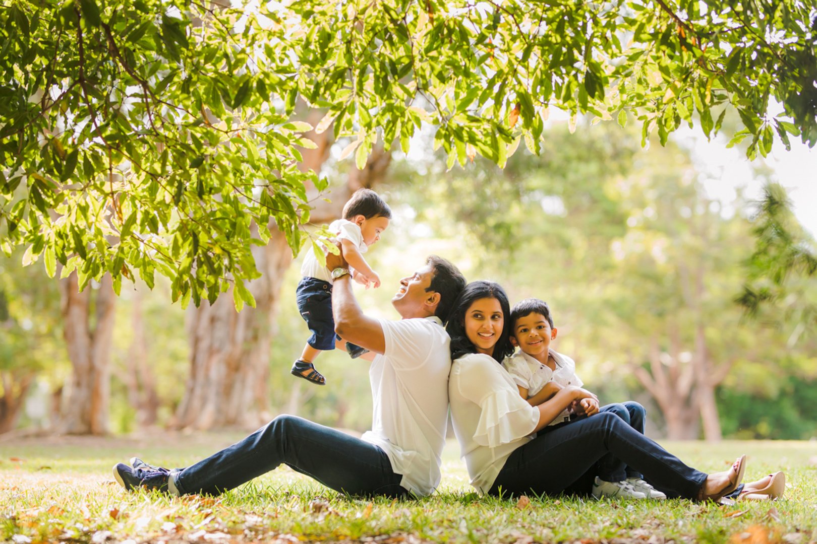 A family of four, sitting on grass under trees, enjoying an outdoor moment. The father, sitting on the grass, is lifting a young boy up. The mother, sitting back-to-back with the father, holds a young girl in her lap. All are smiling, surrounded by g