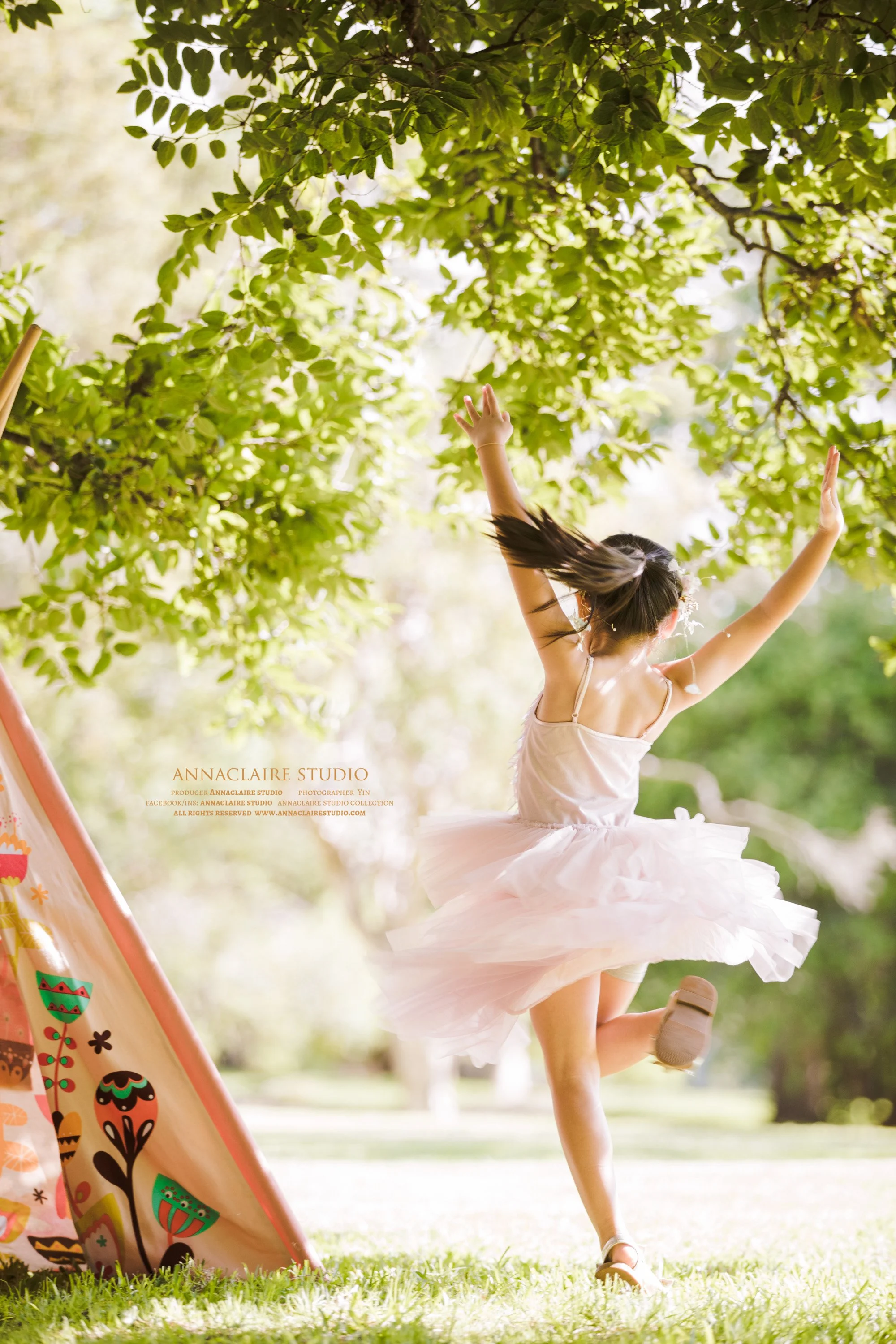 A young girl in a white dress spinning and dancing outdoors under a tree with green leaves, with a colorful tent in the foreground.