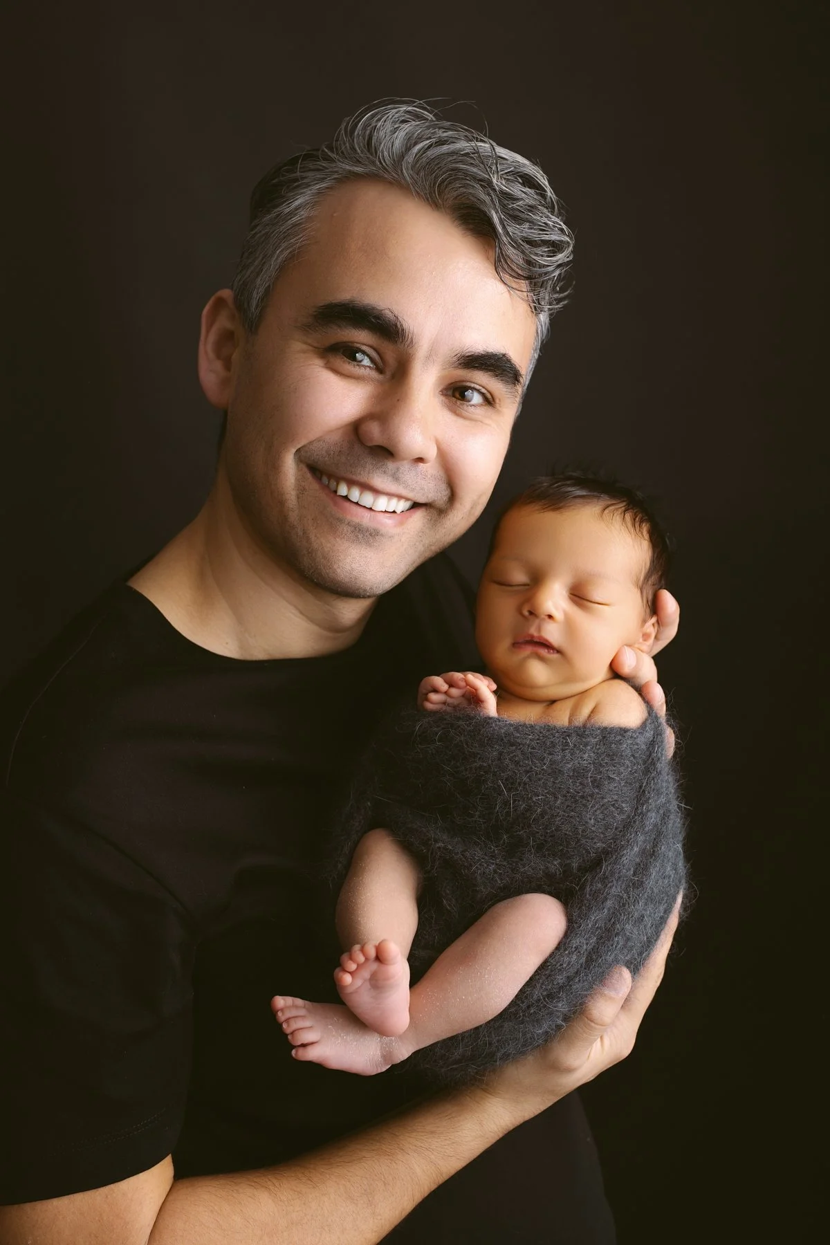 A man with gray hair smiling and holding a sleeping baby wrapped in a dark gray blanket against a dark background.