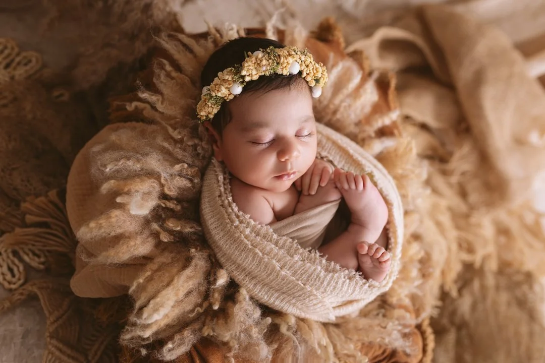 A sleeping baby wearing a floral headband, wrapped in beige cloth, lying on a textured, soft blanket.