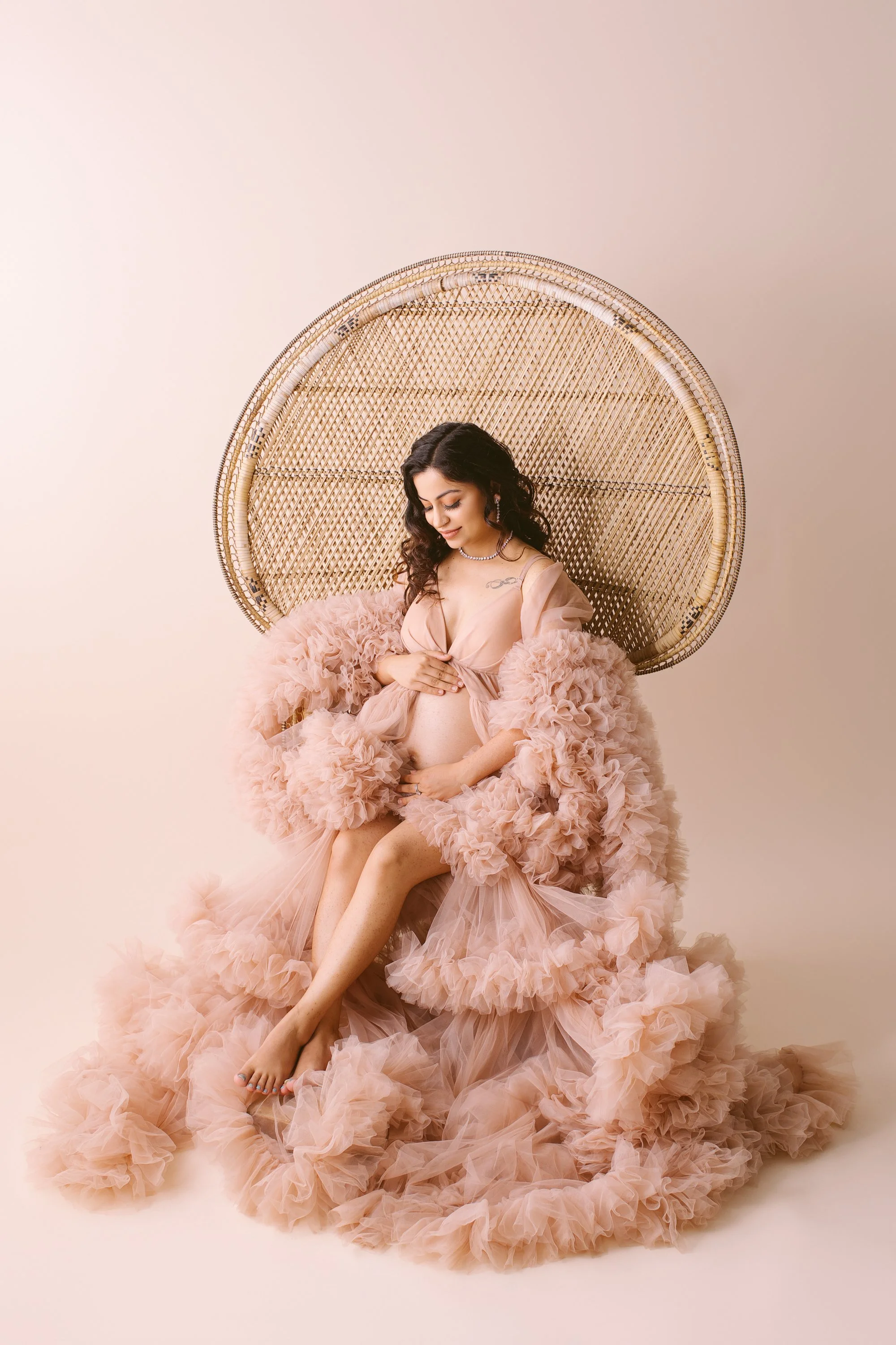 A pregnant woman sitting on a rattan peacock chair, wearing a flowing pink ruffled gown, with her hand on her belly and a gentle smile.