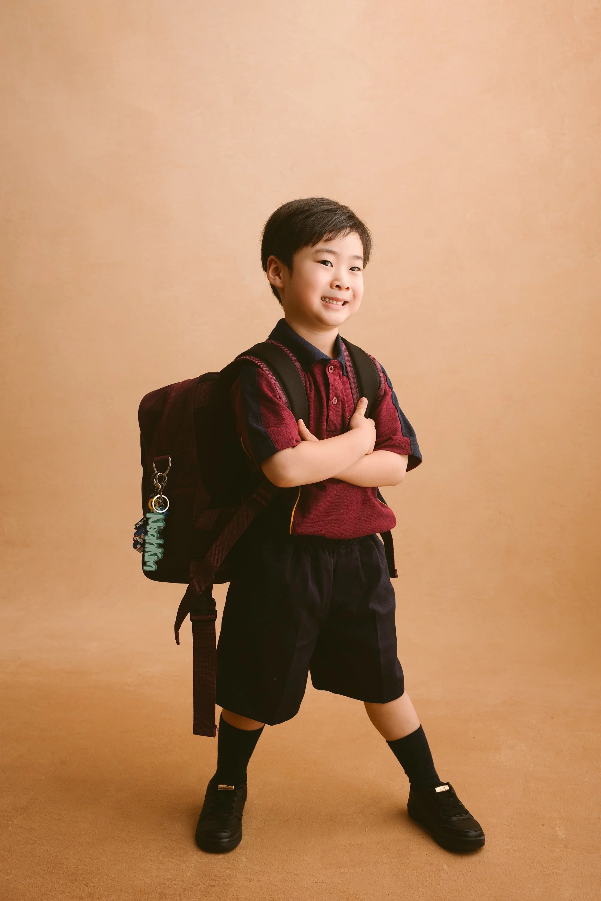 A young boy with short black hair stands with arms crossed, wearing a maroon and navy polo shirt, black shorts, black shoes, and a backpack with a keychain that has the name 'Nashawn', against a beige background.