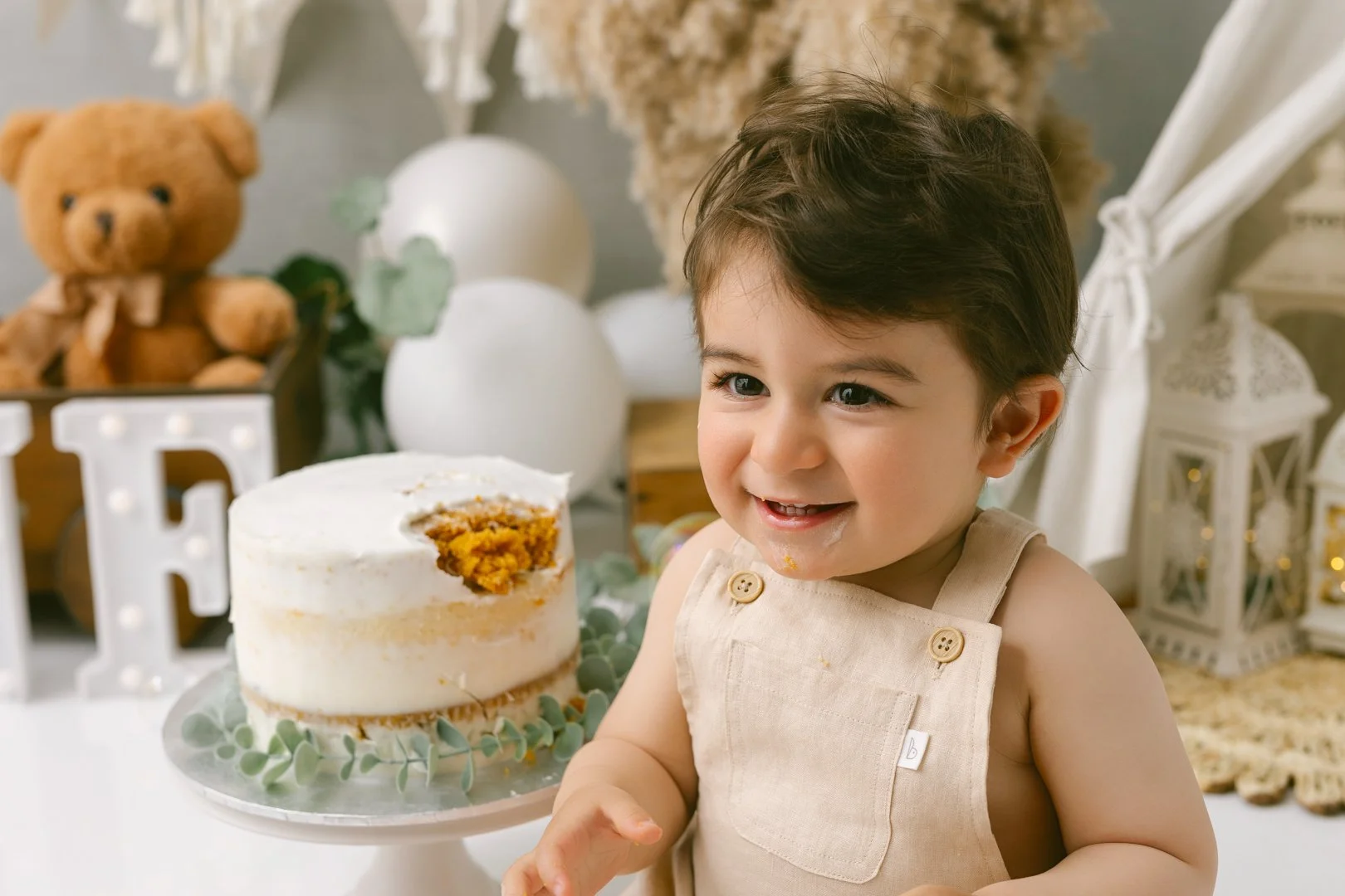 A smiling A  one year old baby with dark hair and wearing a beige sleeveless shirt with buttons, sitting next to a birthday cake with a slice missing, decorated with white frosting and green leaves underneath, on a white cake stand.
