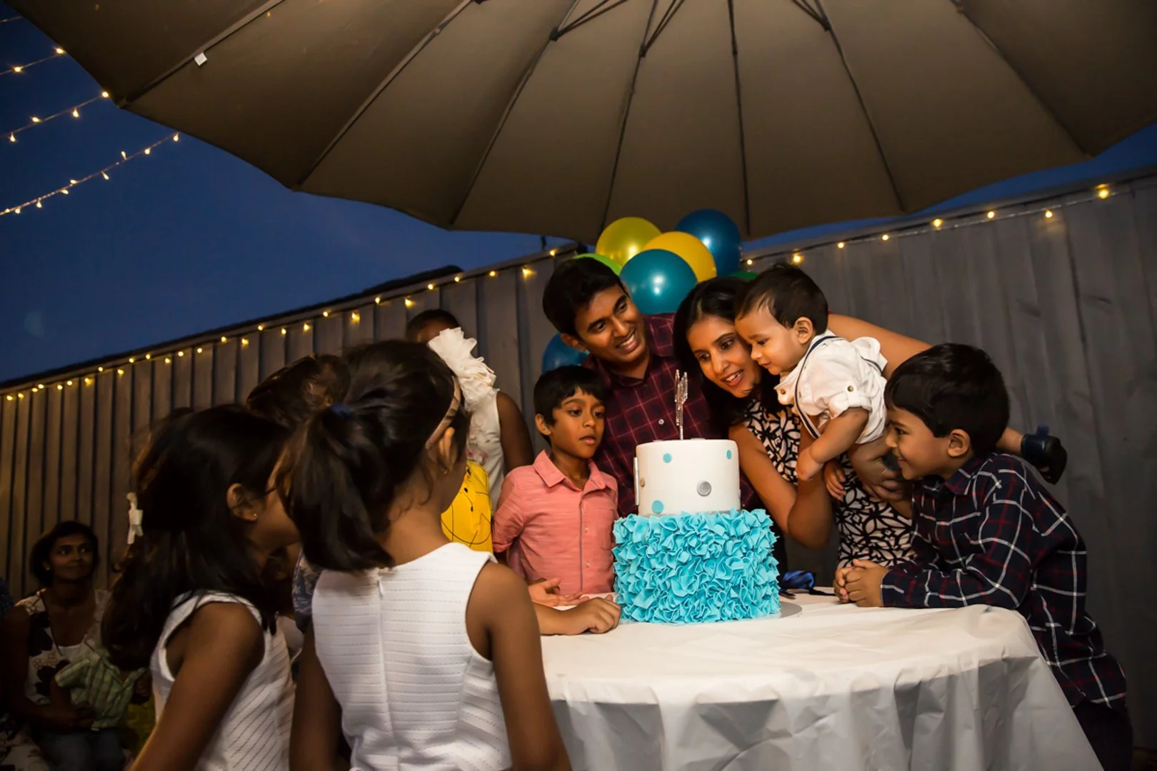 Children and adults gather around a birthday cake outdoors under a large umbrella at night, celebrating a birthday party.